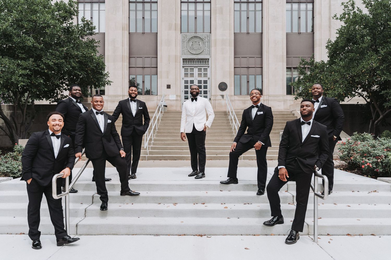 Groomsmen in black tuxedos strike dynamic poses on formal building steps.