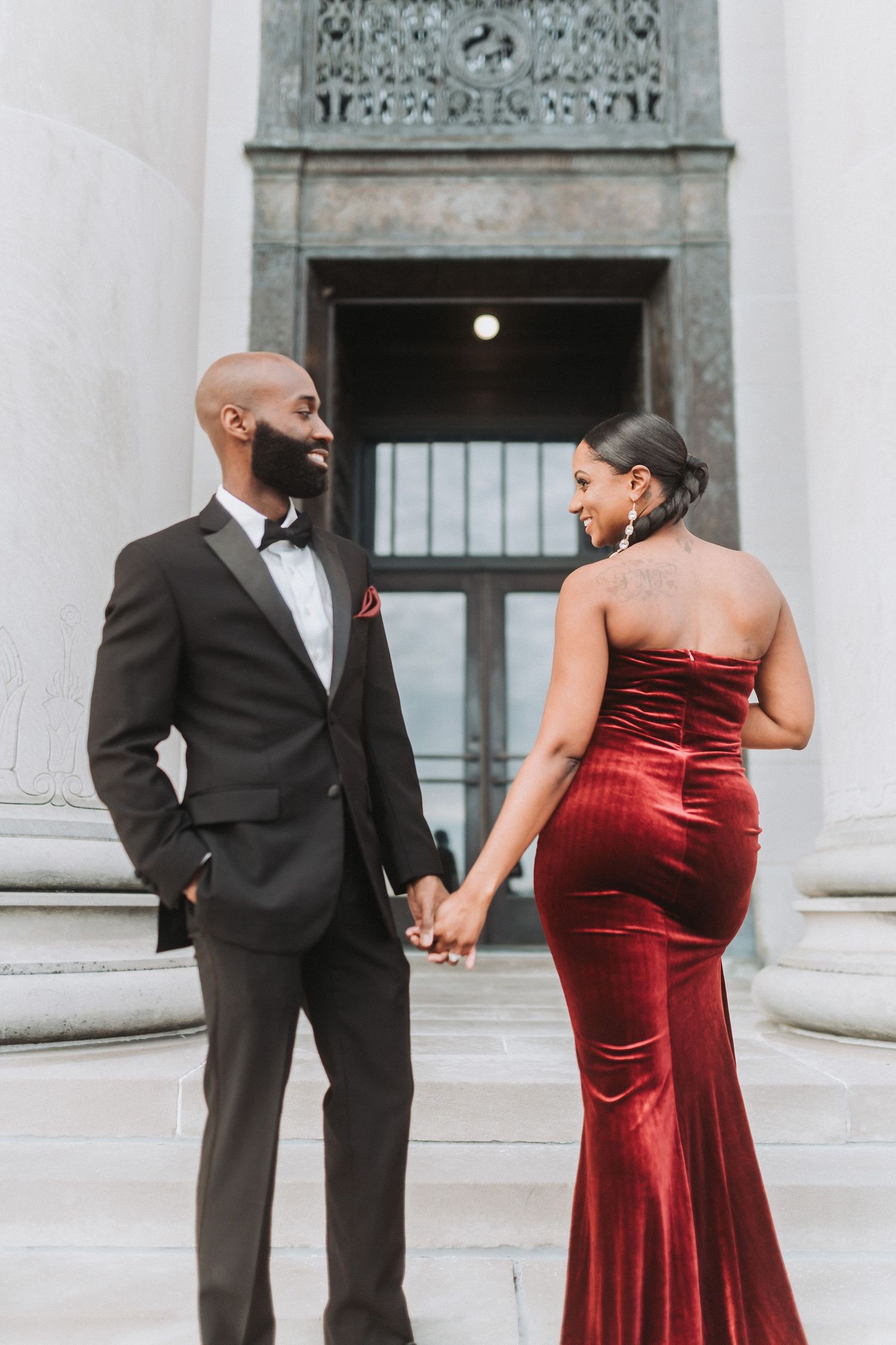 Formal couple in a black tuxedo and red velvet gown hold hands while standing by classical architecture.