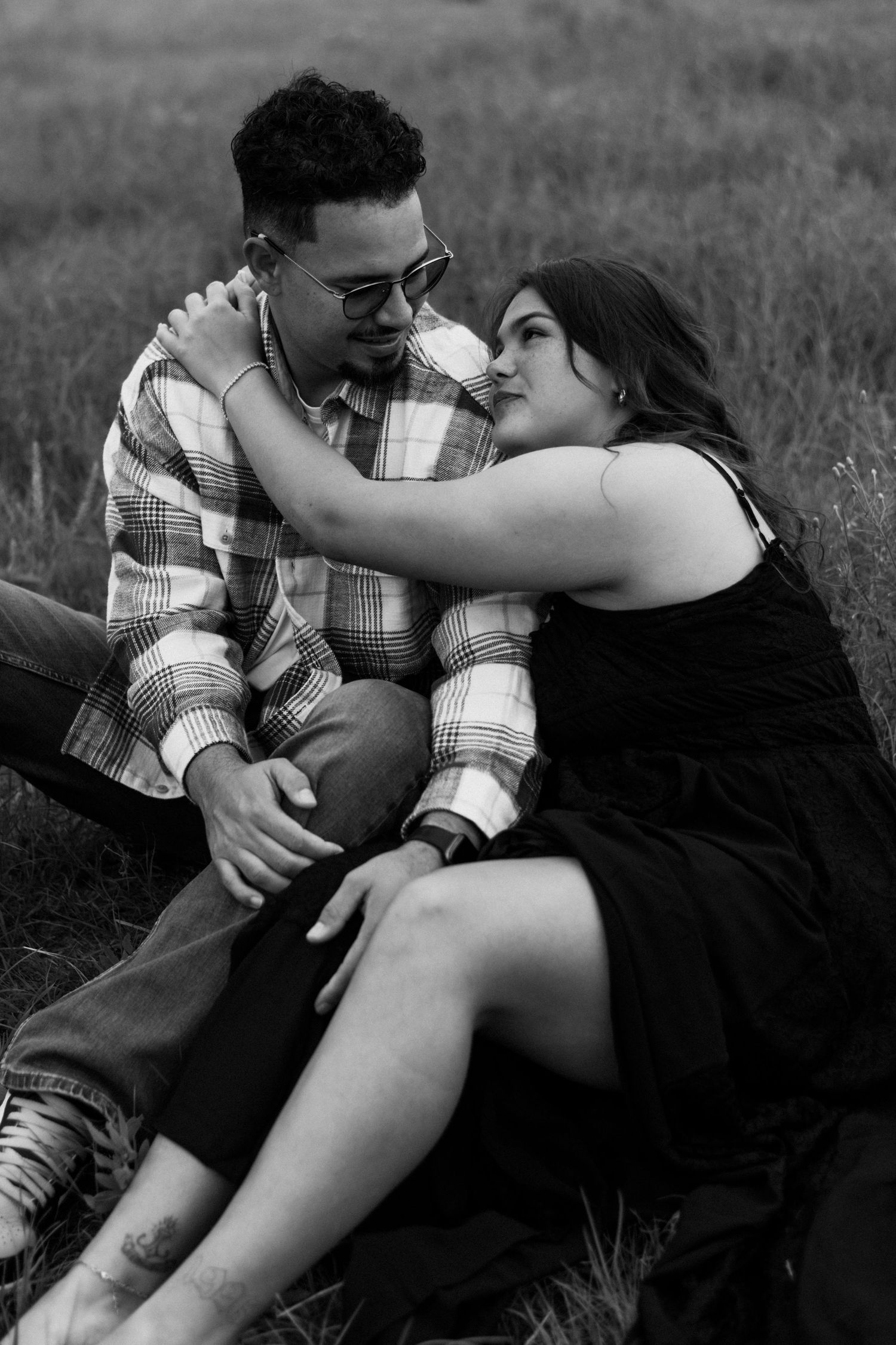 Black and white photo of a couple sitting in a grassy field, sharing a tender romantic moment together.