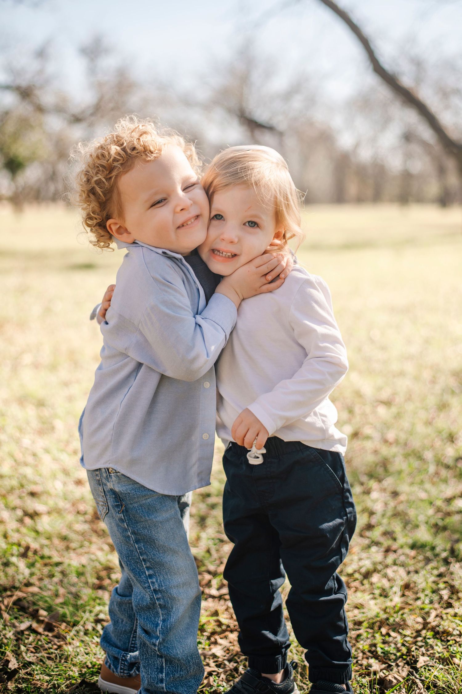 Two young children hugging outdoors in a sunny park, smiling warmly in casual clothes on a bright day.