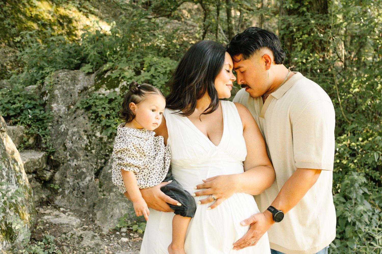 Pregnant woman in white dress with husband and toddler daughter posing outdoors in a lush green forest setting.