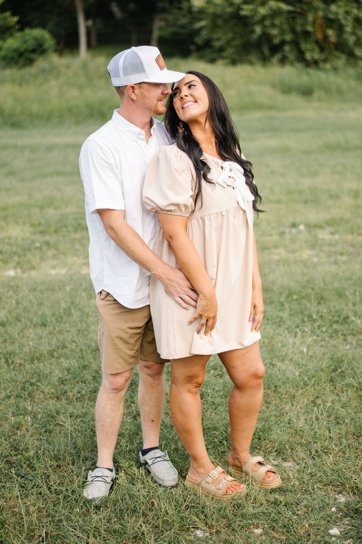 A couple shares a romantic moment in a green field, woman in beige dress smiling up at man in white shirt and cap.