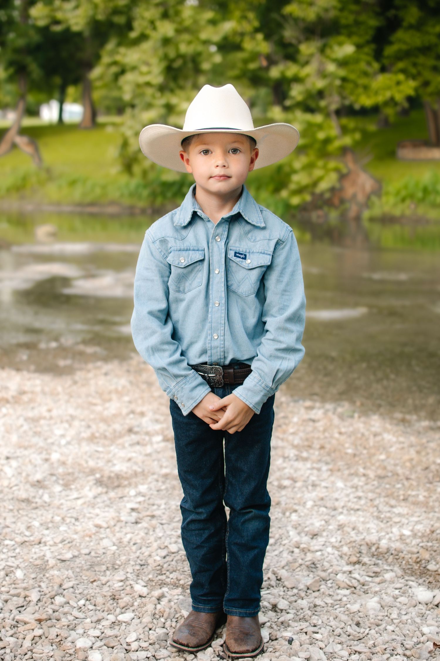 Young boy in denim shirt, cowboy hat, and boots posing near a river on rocky ground.