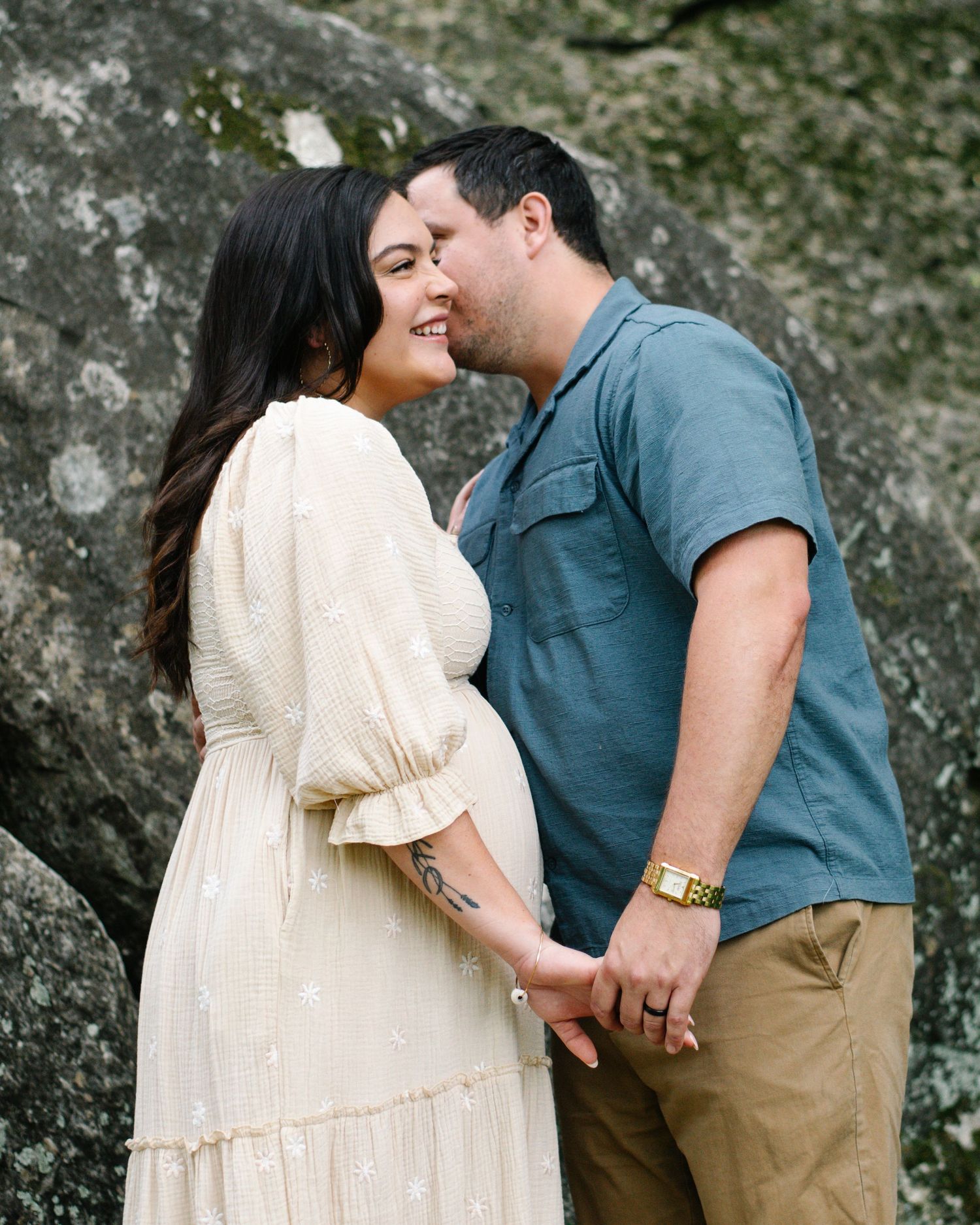 Smiling pregnant woman in white dress holds hands with man in blue shirt as he kisses her cheek outdoors.