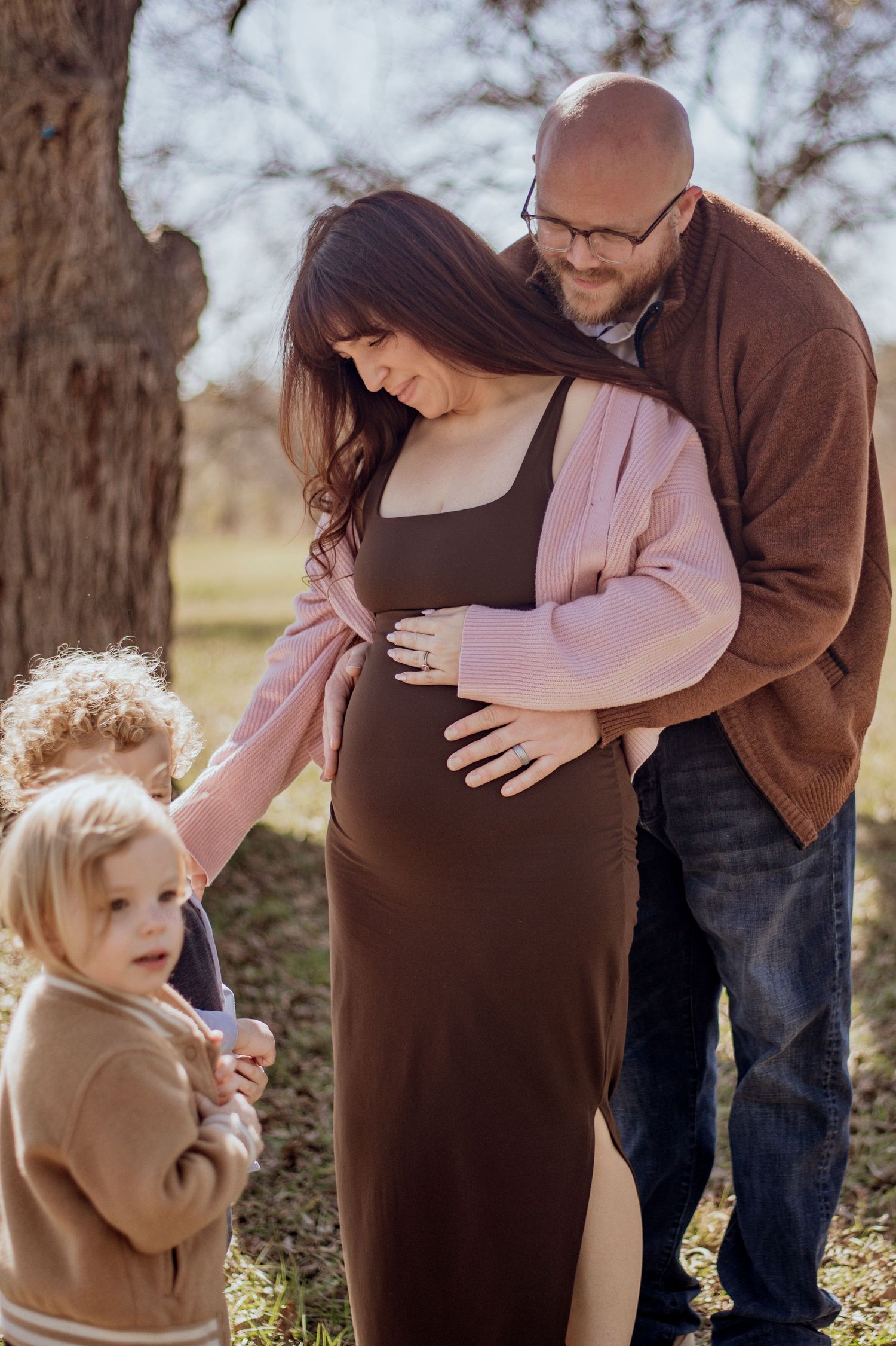 Pregnant woman in brown dress embraced by husband outdoors while two young children touch her baby bump.
