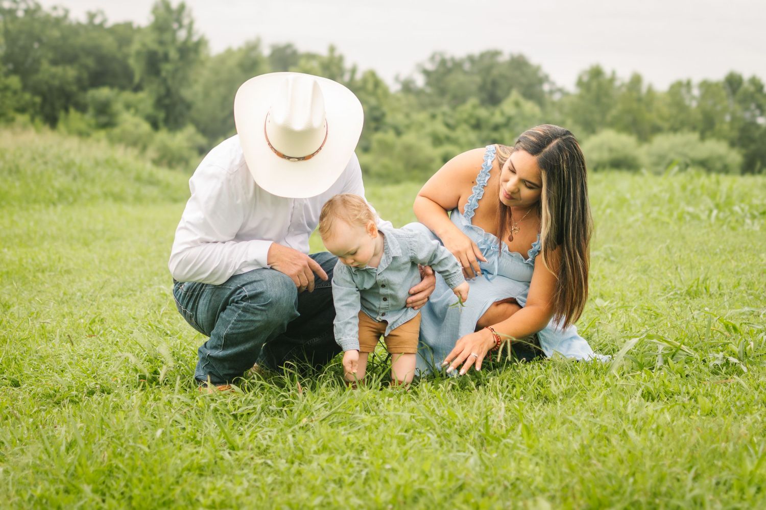 Young family with baby enjoys a sunny day in a lush green meadow, parents smiling as toddler explores the grass.