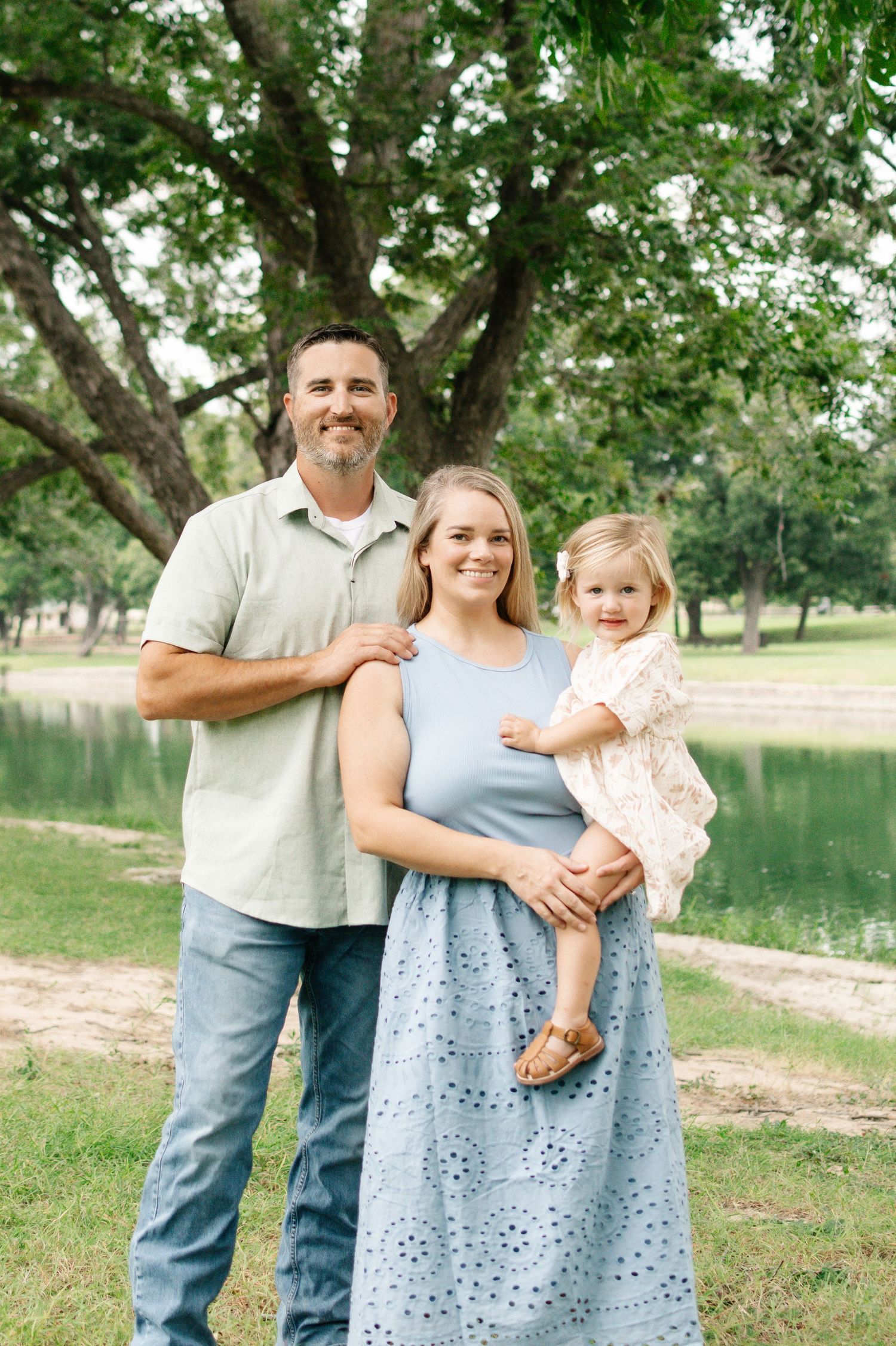 A smiling family of three poses outdoors near a pond, with a man, woman, and toddler girl in a lush green park.