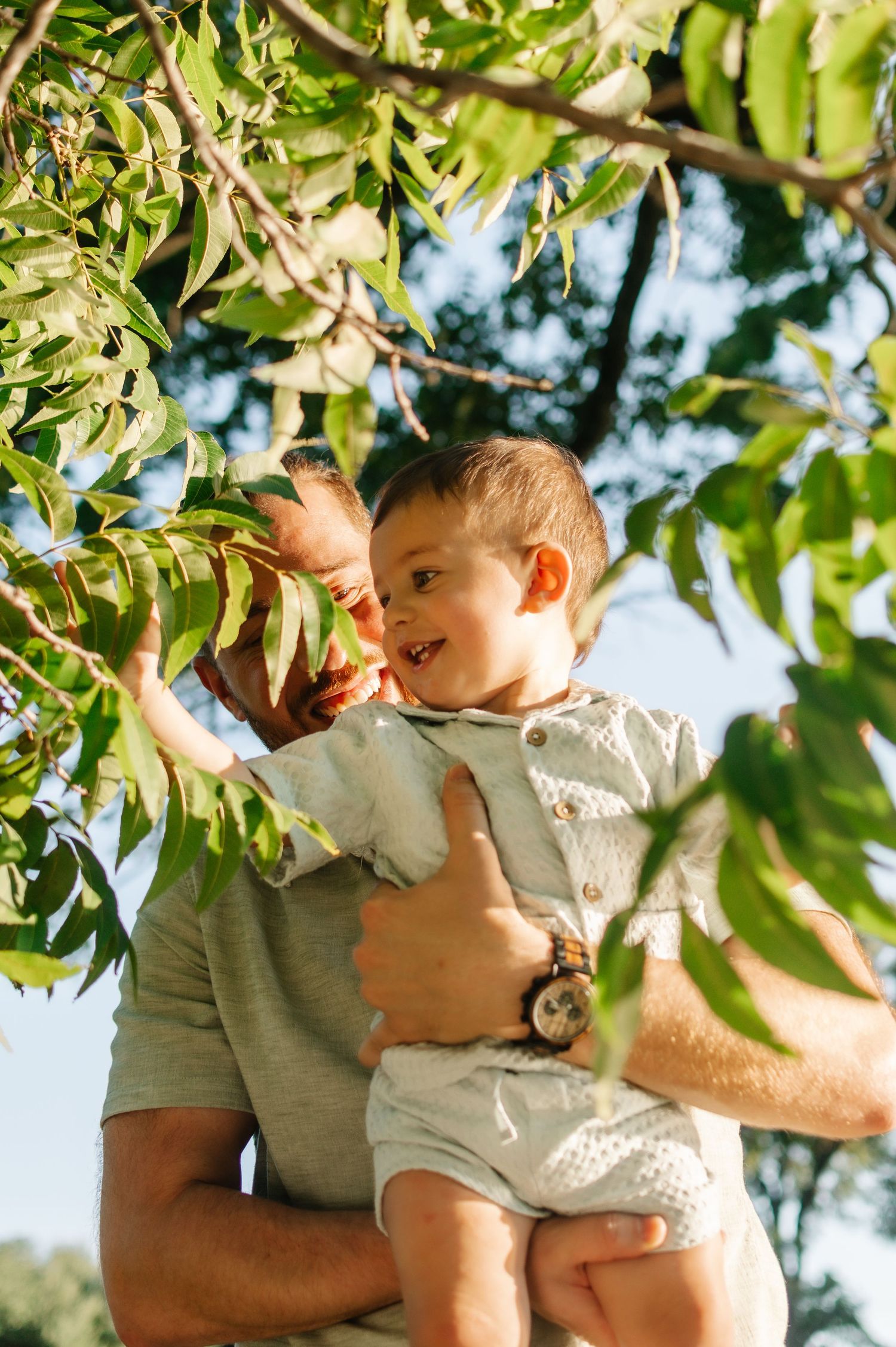 A man holds a smiling toddler up to touch green leaves on a tree branch outdoors in warm sunlight.