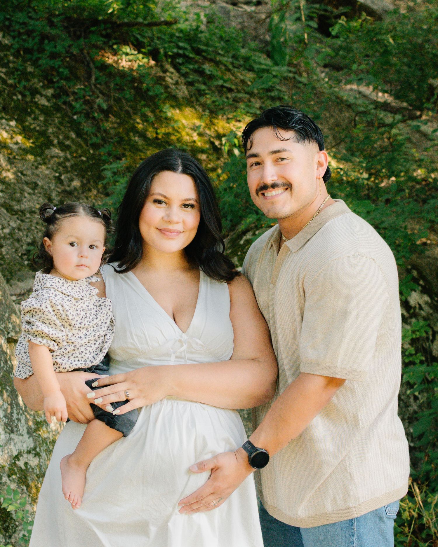 A happy family of three poses outdoors, with a pregnant woman in white dress, man in beige shirt, and baby girl.