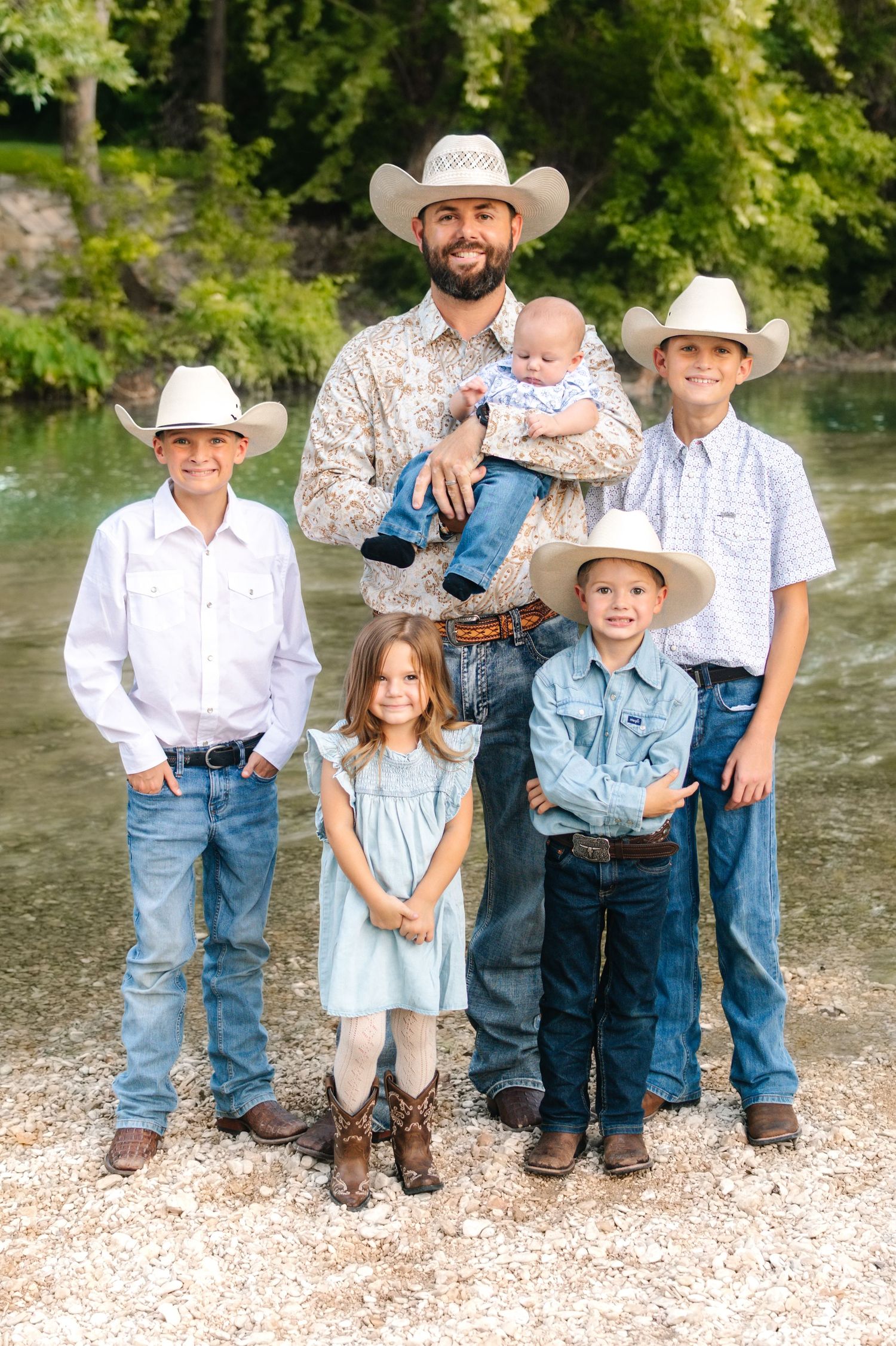 Father in cowboy hat holds baby surrounded by four children in western wear by a river.