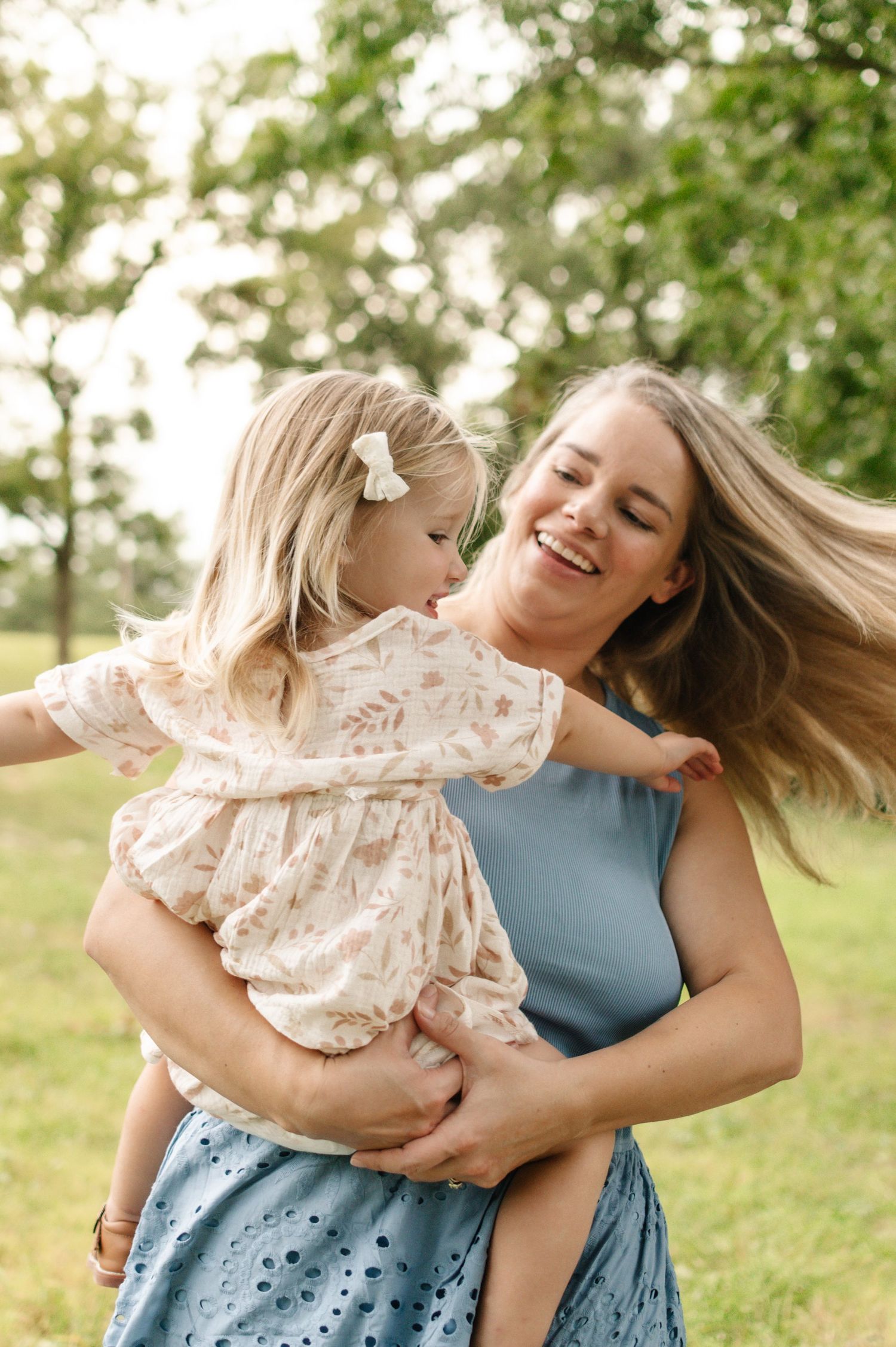 A joyful mother holds her laughing blonde toddler daughter outdoors in a lush green park setting.
