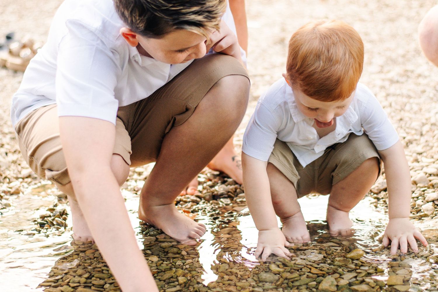 Adult and red-haired toddler crouching together, exploring shallow rocky stream water outdoors.