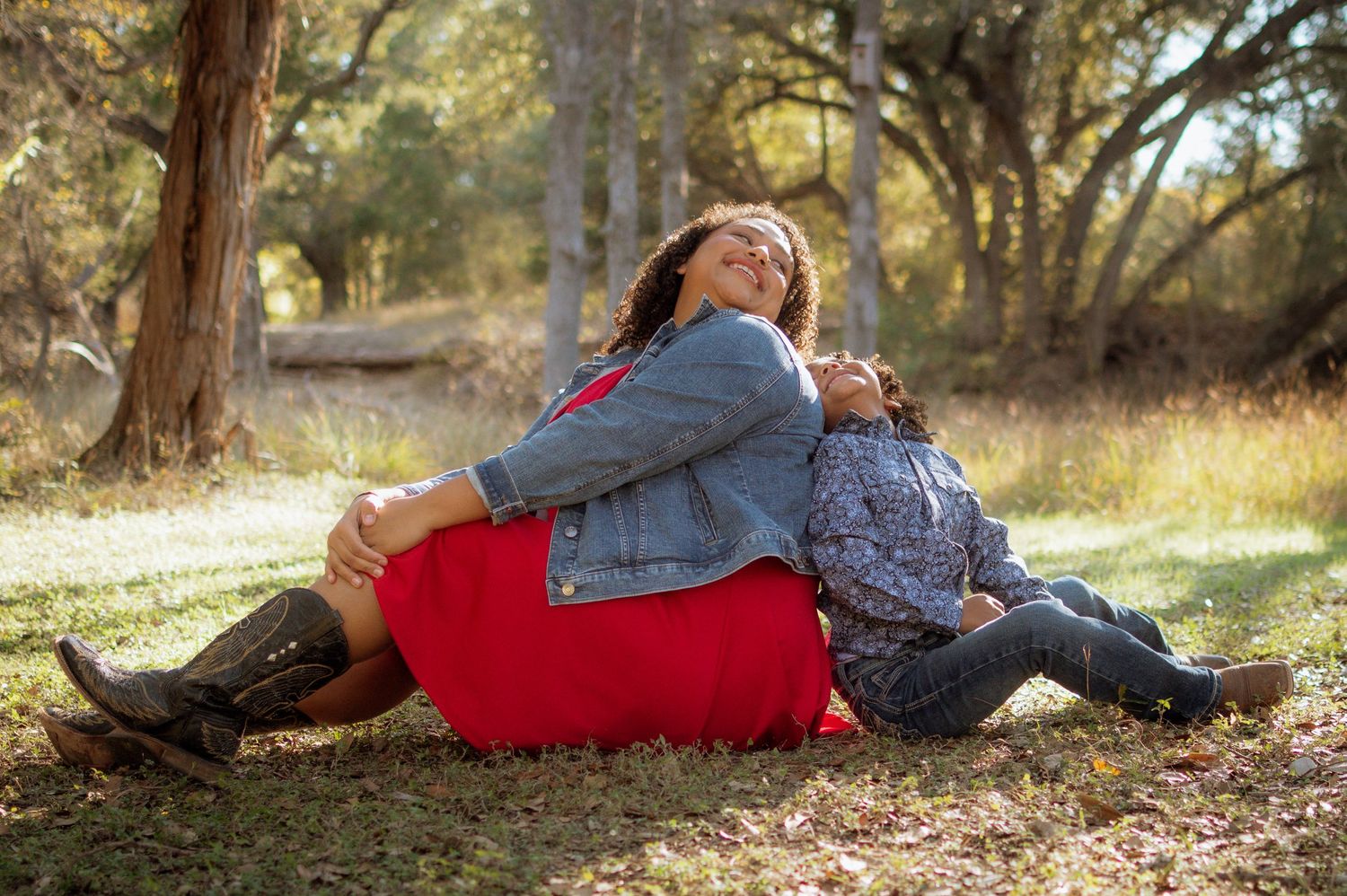 Two women sitting back-to-back on grass in autumn park, one in red dress and denim jacket, smiling joyfully.