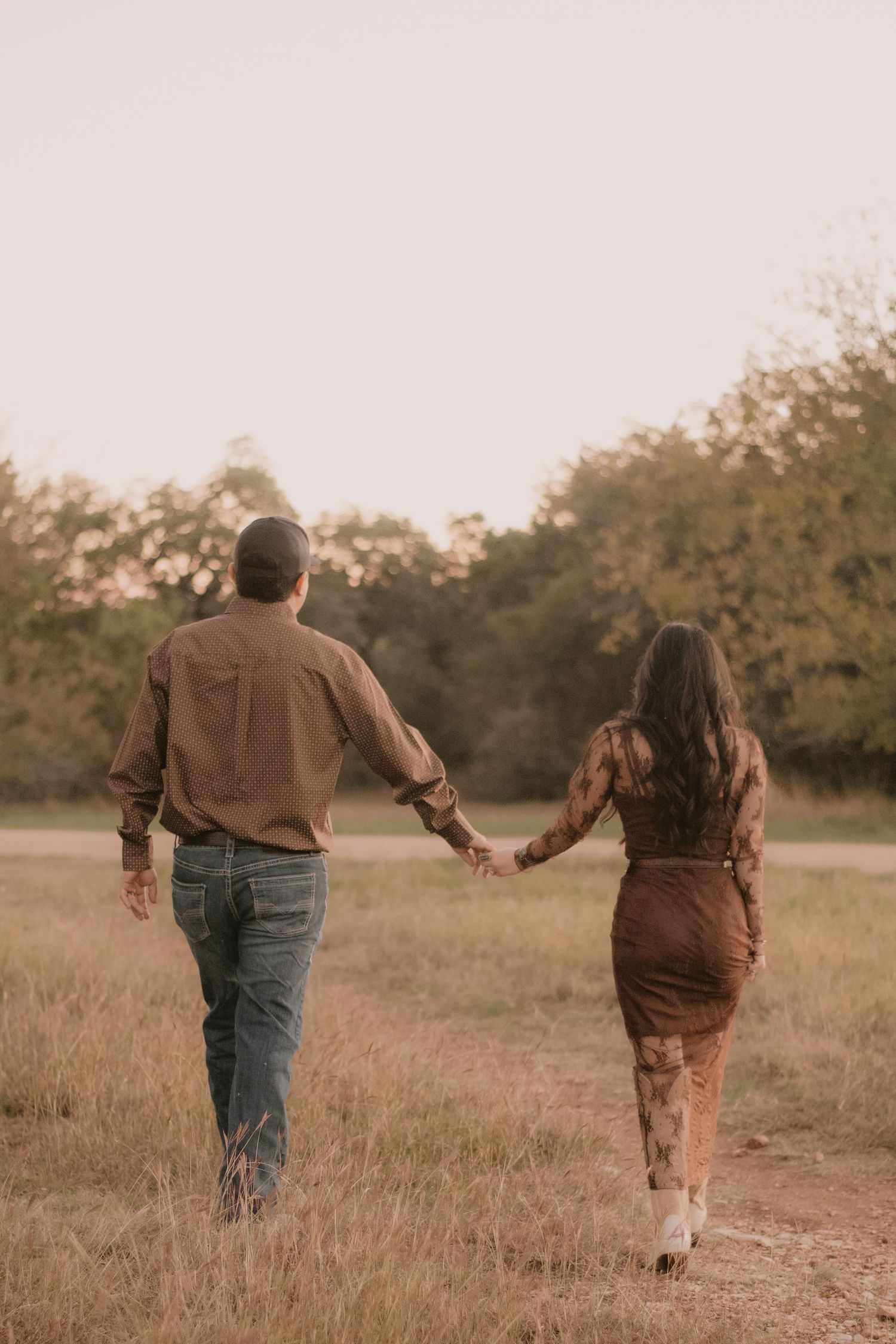 A couple holds hands walking through a golden field toward trees at sunset, both dressed in earthy brown tones.
