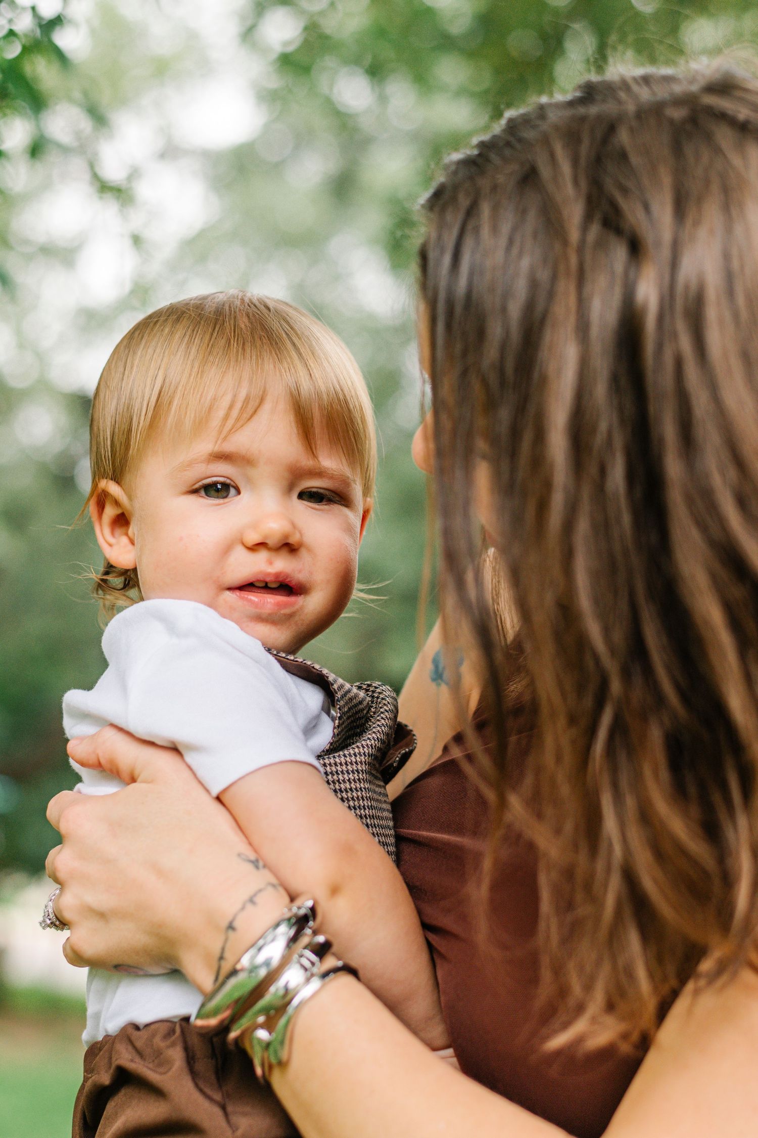 A mother holds her blonde toddler outdoors in a green park, the child looking curiously at the camera.
