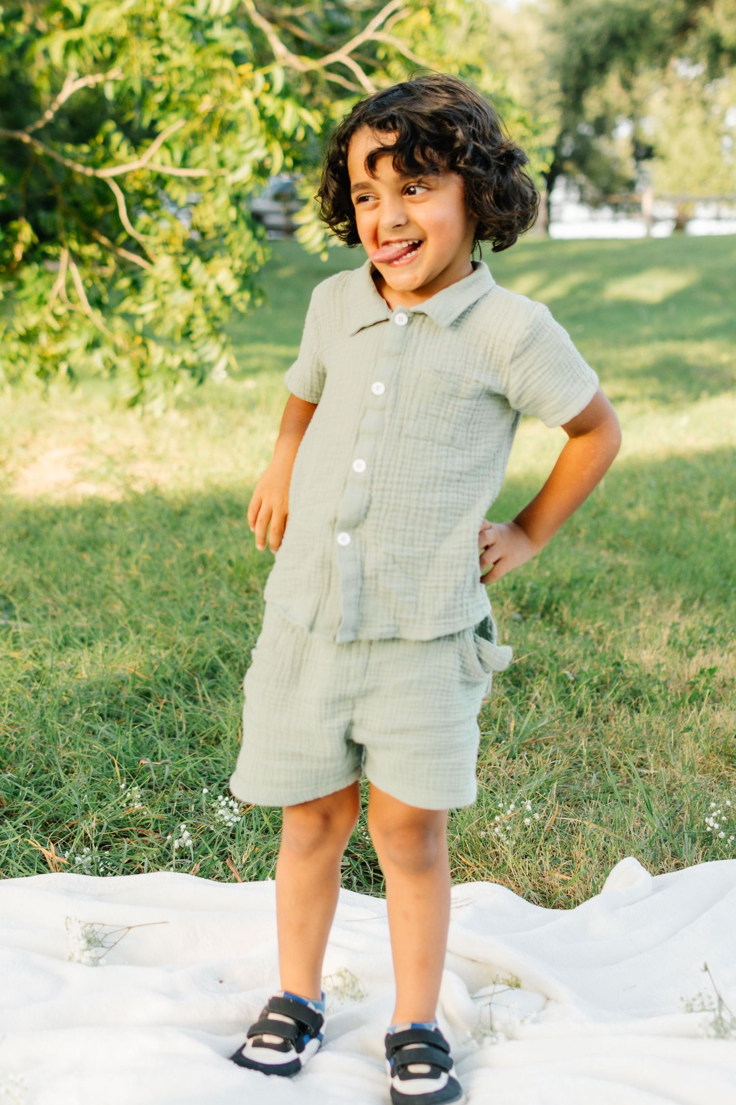 Young boy in sage green linen shorts set smiling outdoors on white blanket in sunny park.