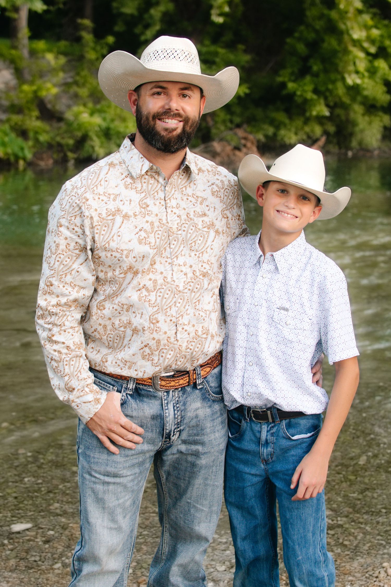 A bearded man and young boy in matching cowboy hats and western shirts pose together near a river.