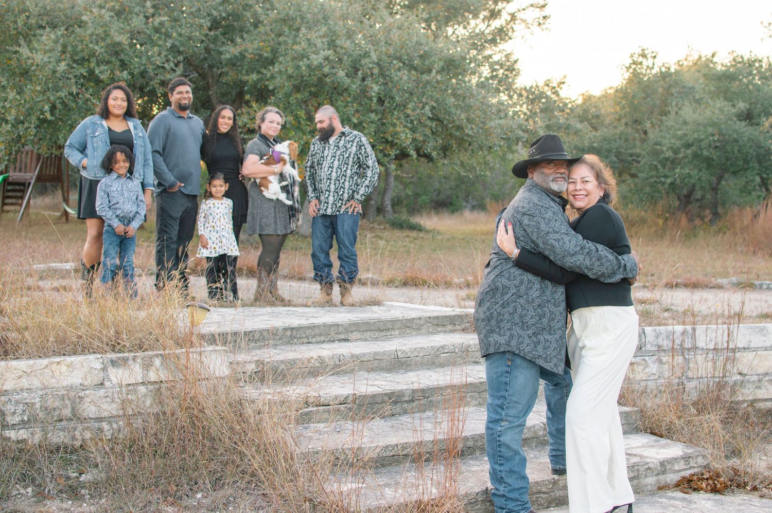 A multigenerational family gathers outdoors on rustic stone steps, with an elderly couple embracing warmly in the foreground.
