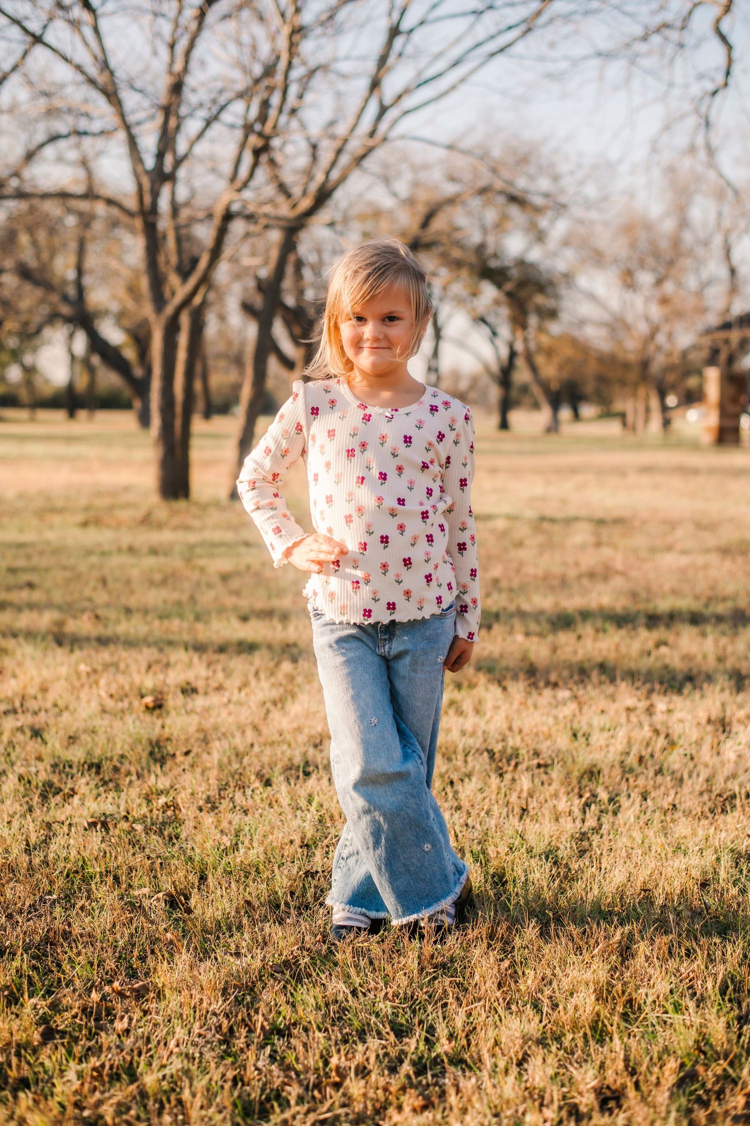 Young girl in floral top and jeans posing in a sunny park with bare trees in the background.