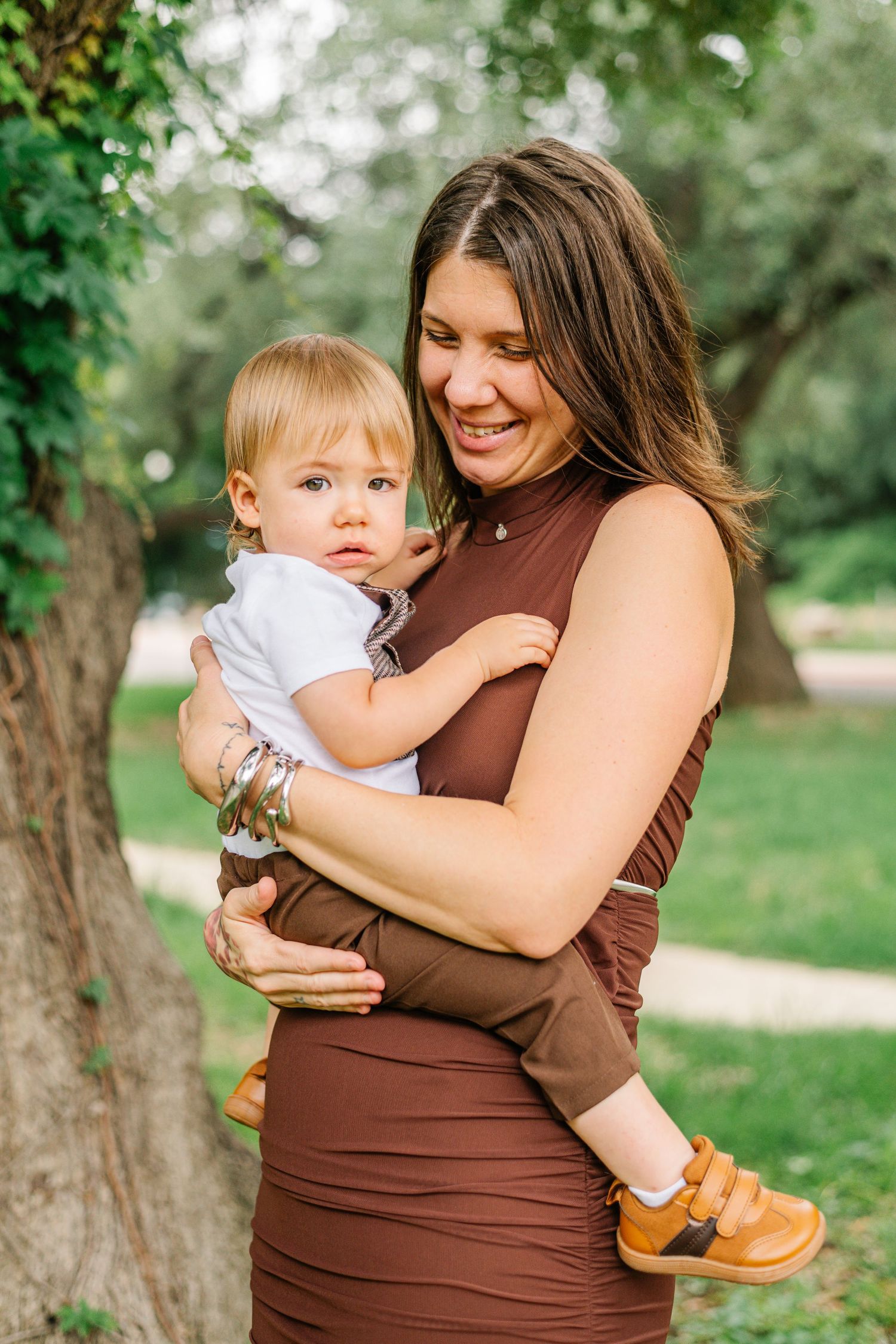 A smiling woman in a brown dress holds a toddler in a white outfit outdoors in a lush green park.