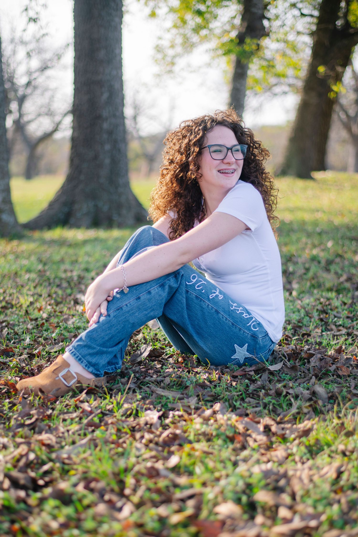 A person in blue jeans and white shirt sits on autumn leaves beneath tall trees in a park setting.