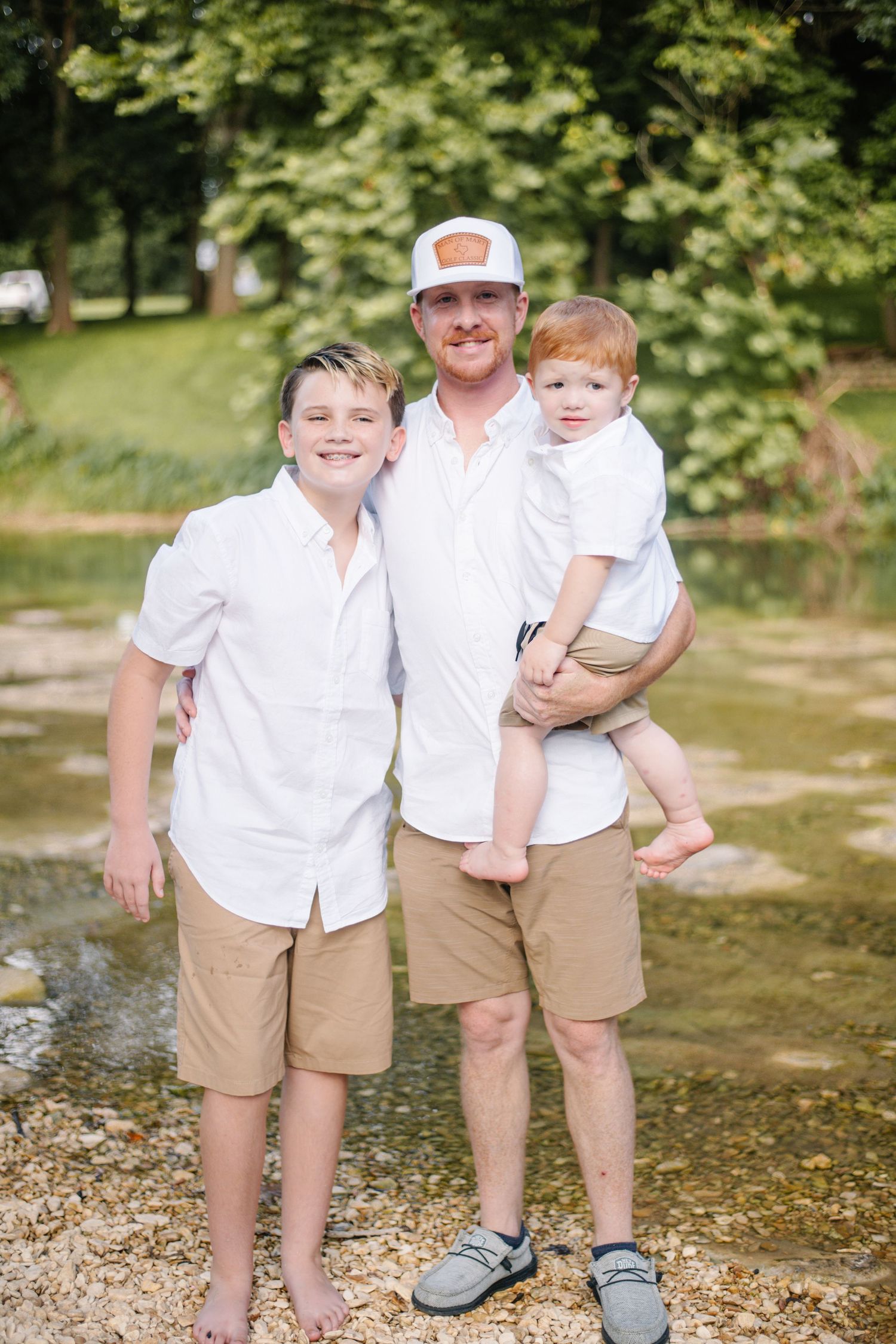 A father in a white shirt and cap poses with two young sons outdoors near a creek, all wearing matching white shirts and tan shorts.
