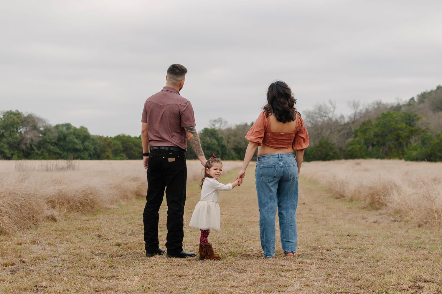 Family of three holding hands in a golden field, parents and toddler viewed from behind on a cloudy day.