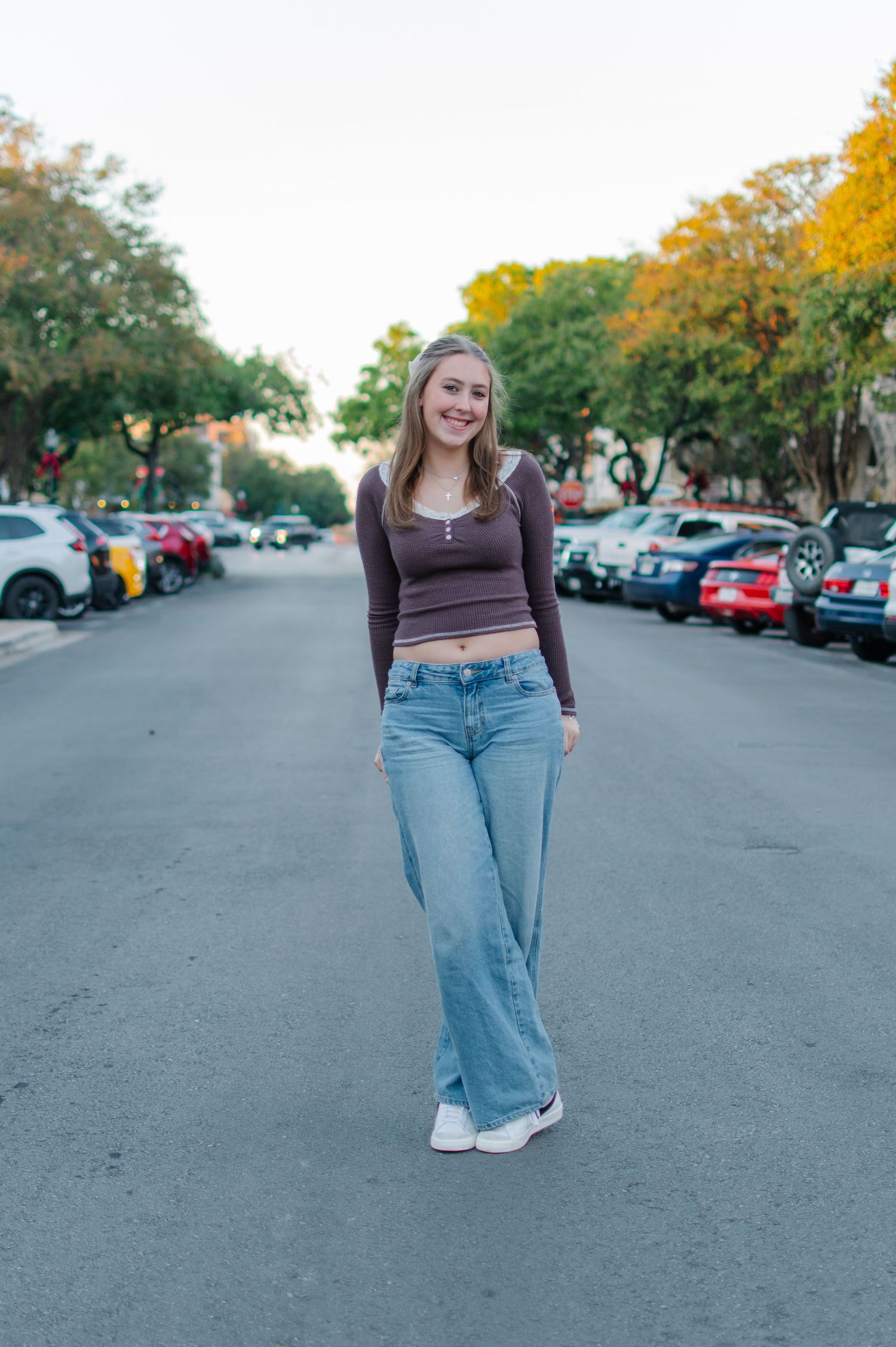 Smiling young woman in brown crop top and wide-leg jeans standing on a tree-lined street.