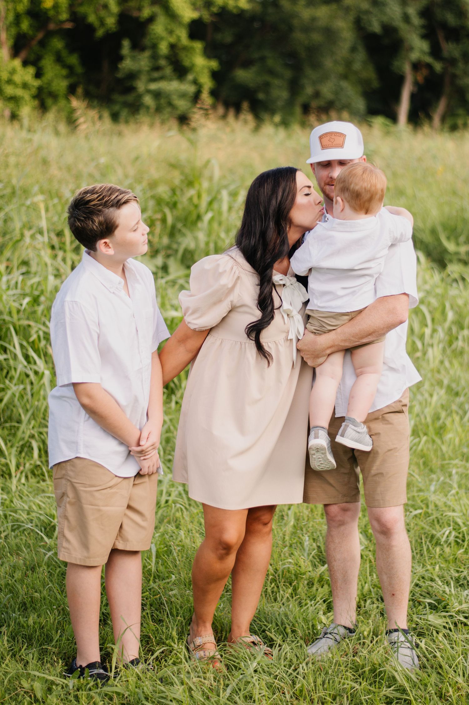 Family of four posing outdoors in matching neutral outfits, mother kissing toddler held by father, older son standing nearby.