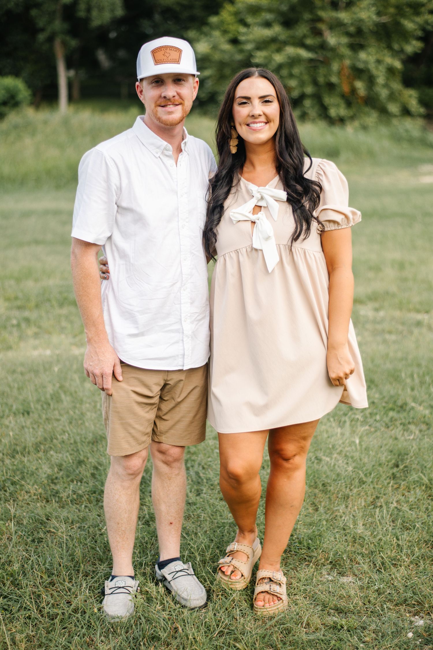 A smiling couple poses outdoors on green grass, man in white shirt and khaki shorts, woman in beige mini dress.