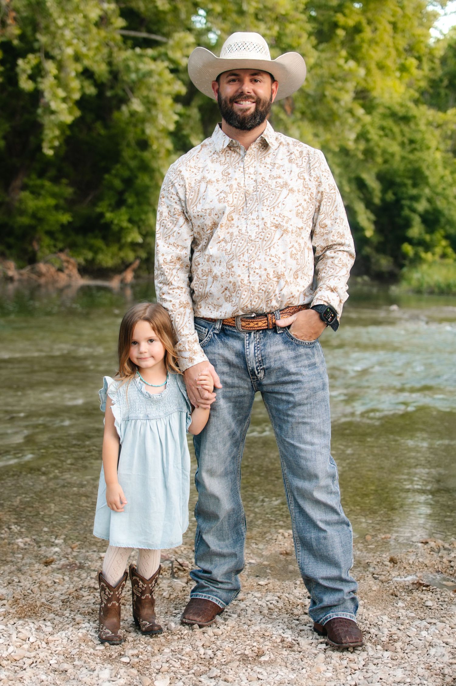A bearded man in a cowboy hat and jeans holds hands with a little girl in a blue dress and cowboy boots outdoors.