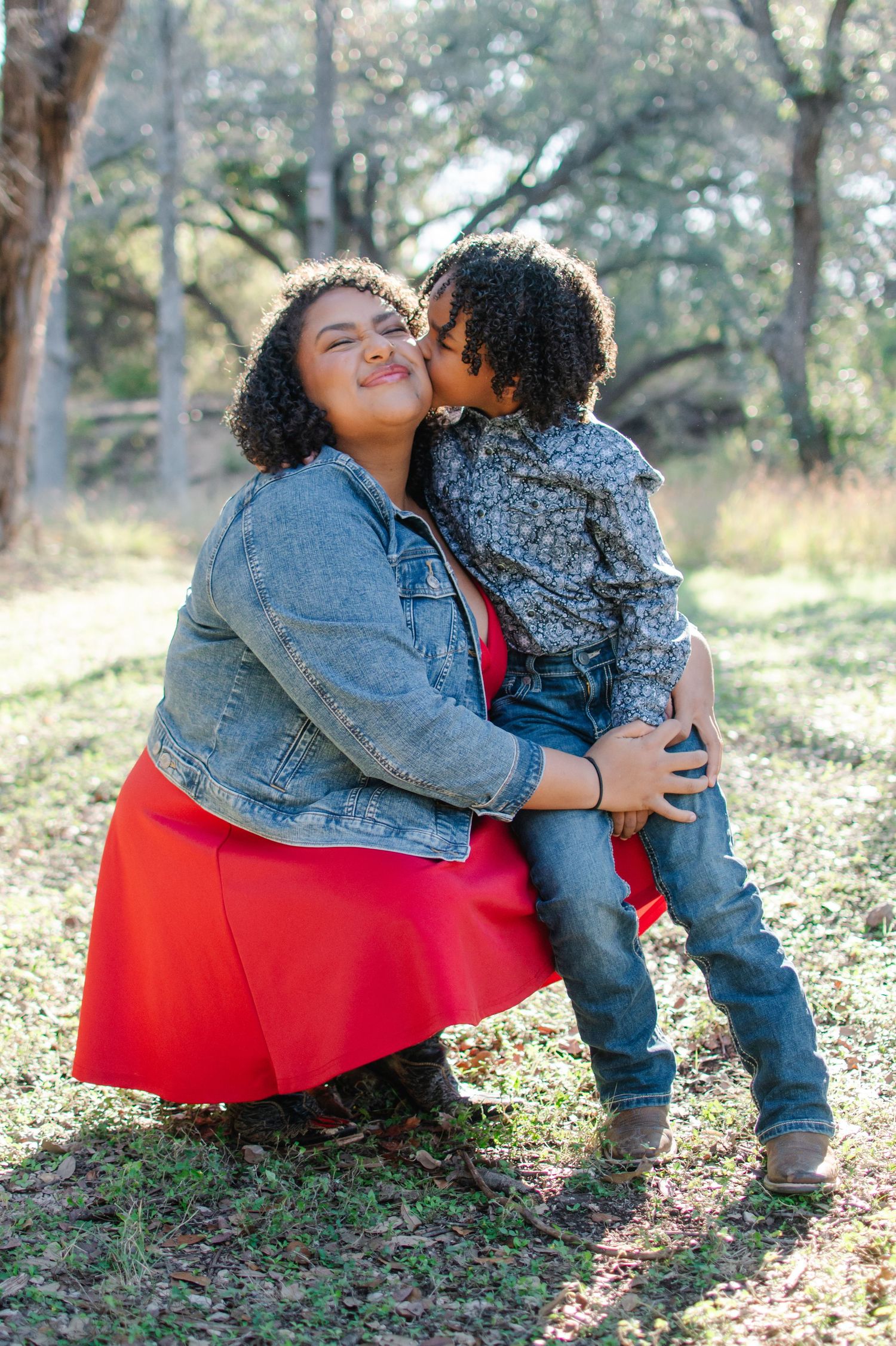 A young child kisses a smiling woman on the cheek outdoors in a sunlit park setting.