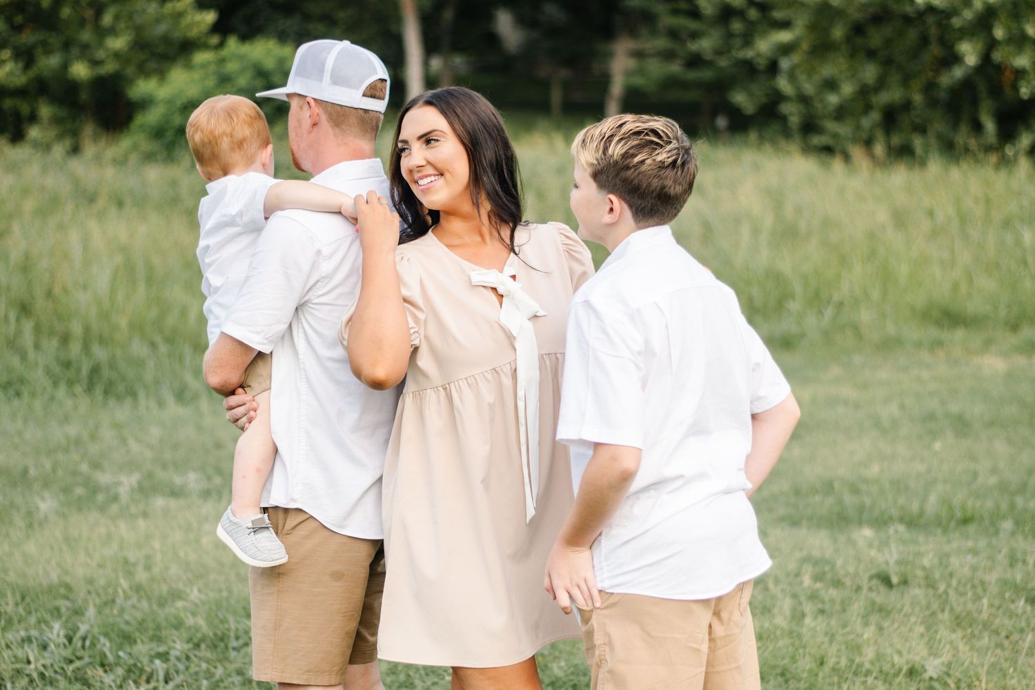 Family of four posing outdoors on green grass, parents with two young boys in coordinating neutral beige and white outfits.