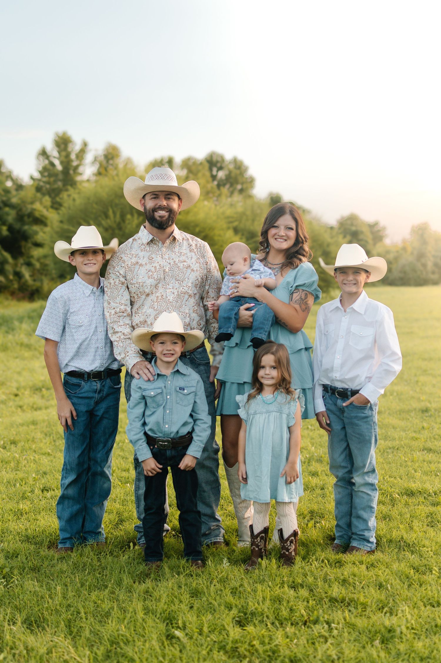 Family of seven in cowboy hats posing in a green field, wearing coordinated western outfits in teal and white.