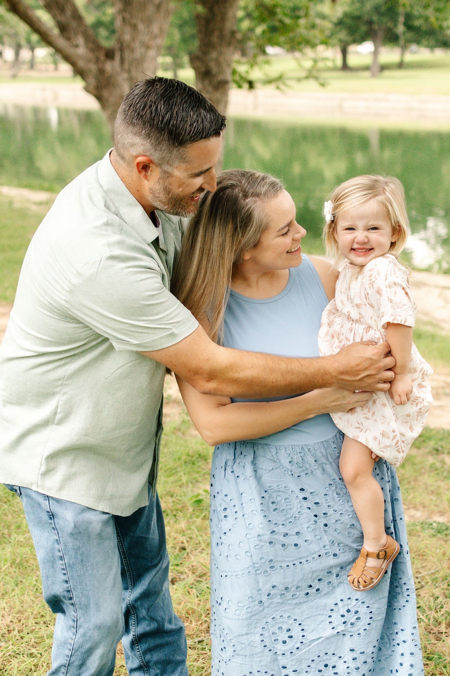 A happy family of three poses outdoors, with parents embracing their smiling toddler daughter in a park setting.