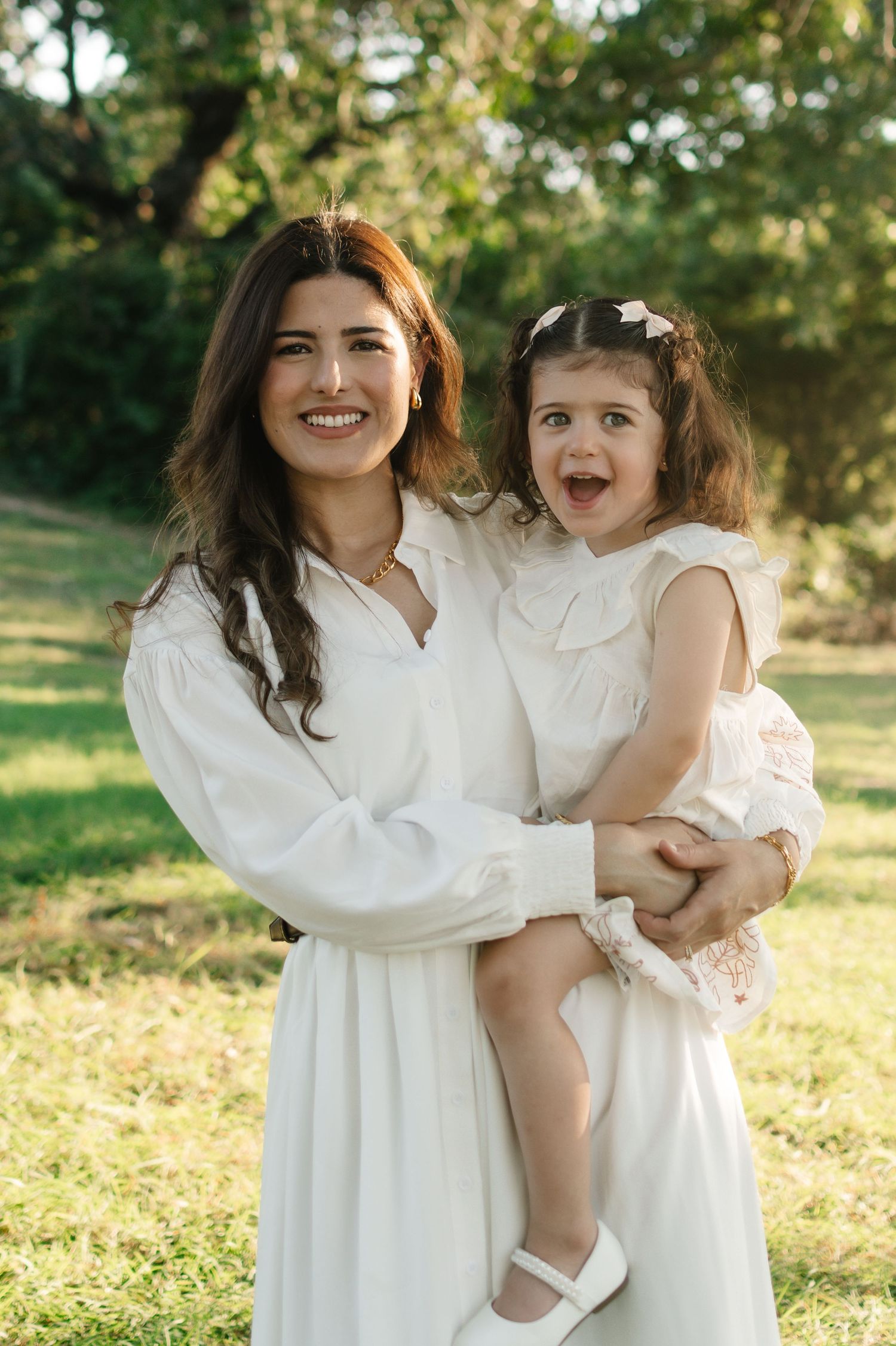 A smiling woman in white dress holds an excited toddler girl outdoors in a sunny green park.