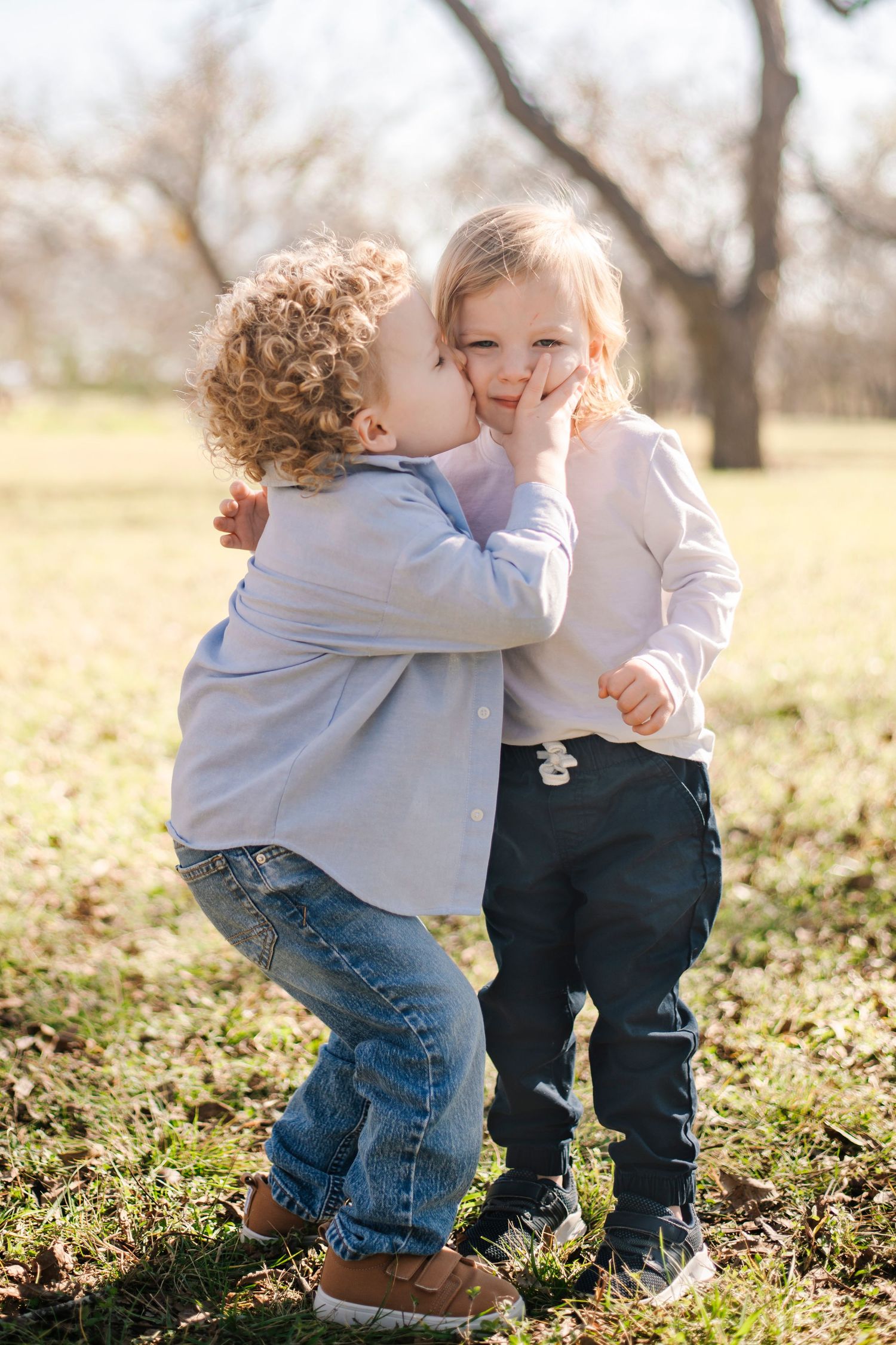 Two young toddlers sharing a sweet kiss on the cheek outdoors in a sunny park setting.