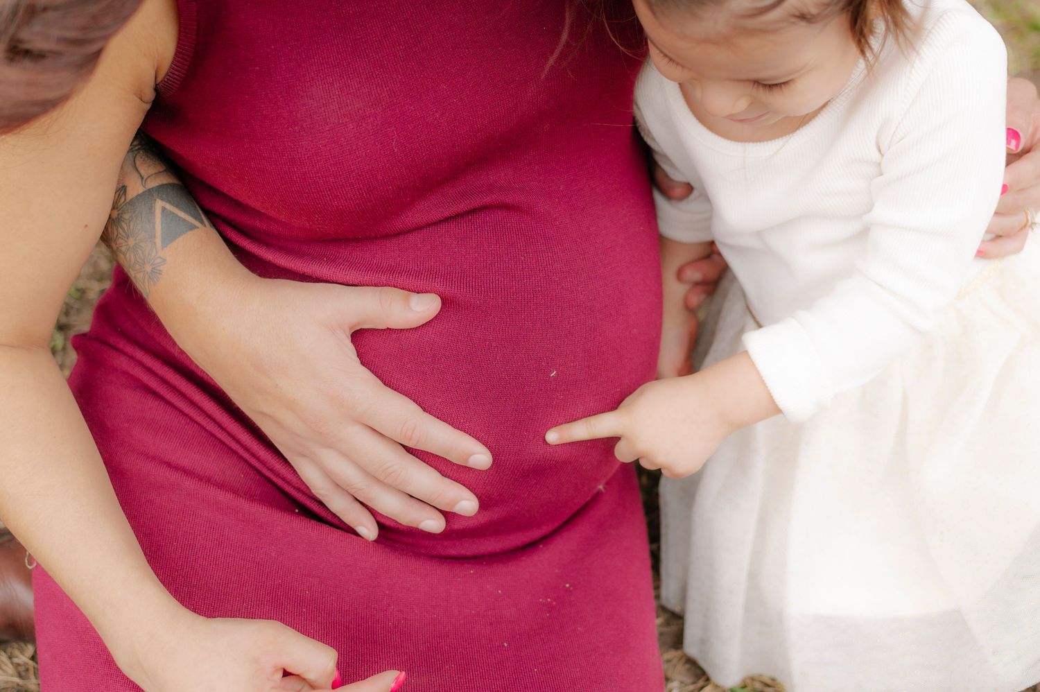 Pregnant woman in red dress with toddler girl touching her baby bump outdoors.
