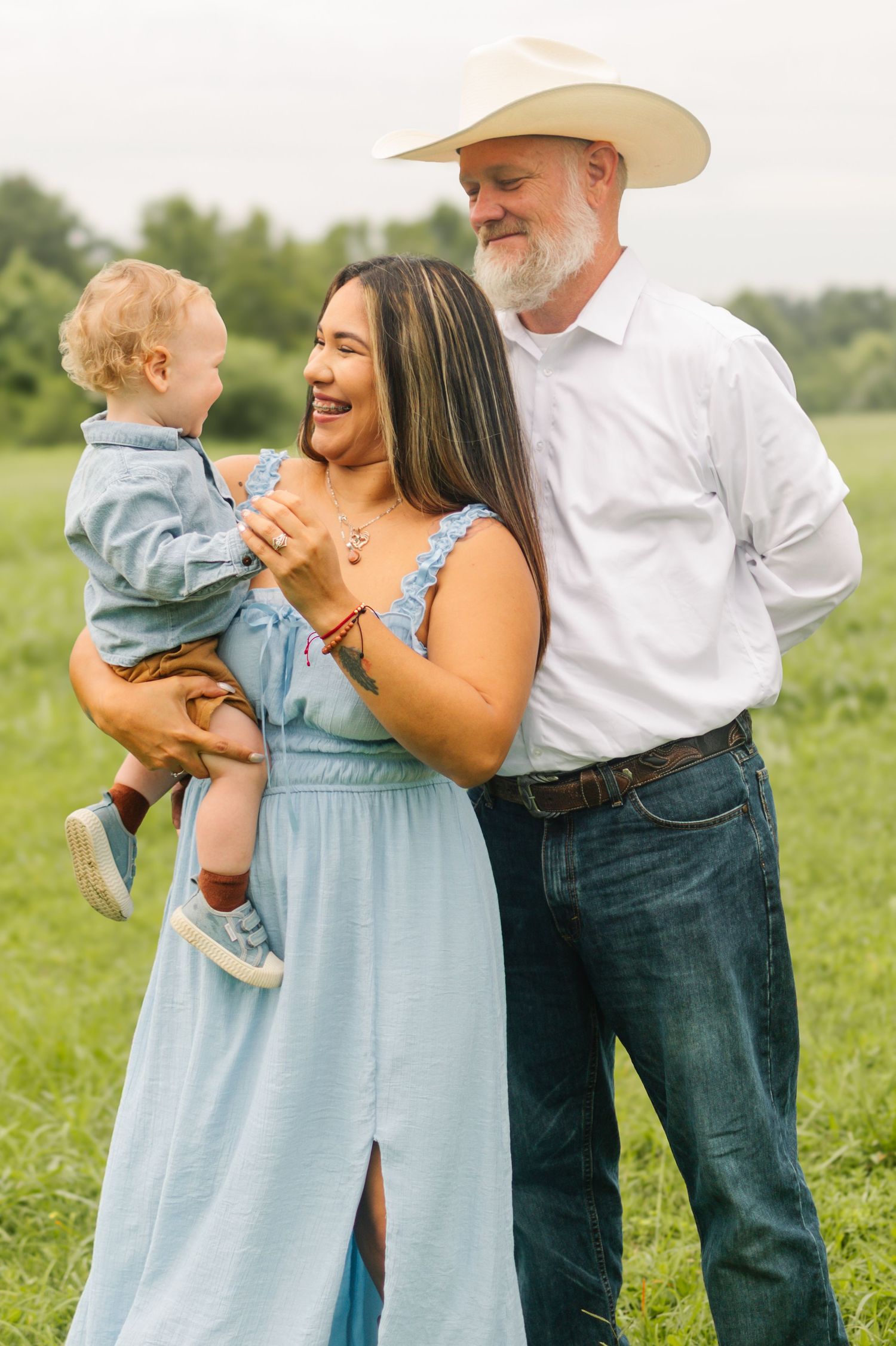 A happy family with a woman in a blue dress, a man in a cowboy hat, and a baby boy posing in a green field.