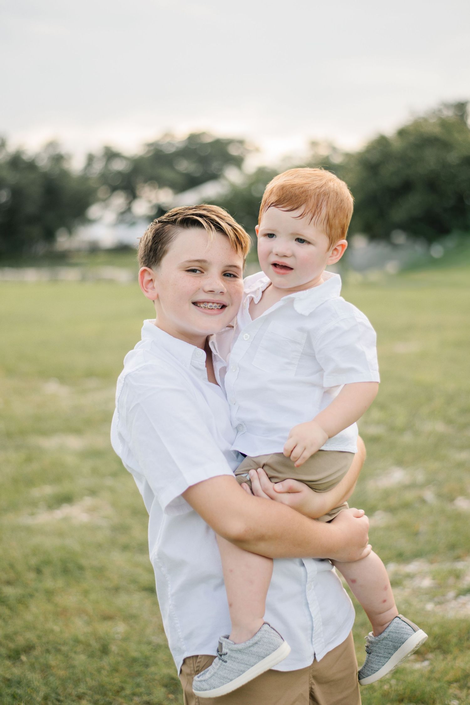 Two brothers in matching white shirts and khaki pants pose outdoors, older boy smiling while holding his red-haired toddler sibling.