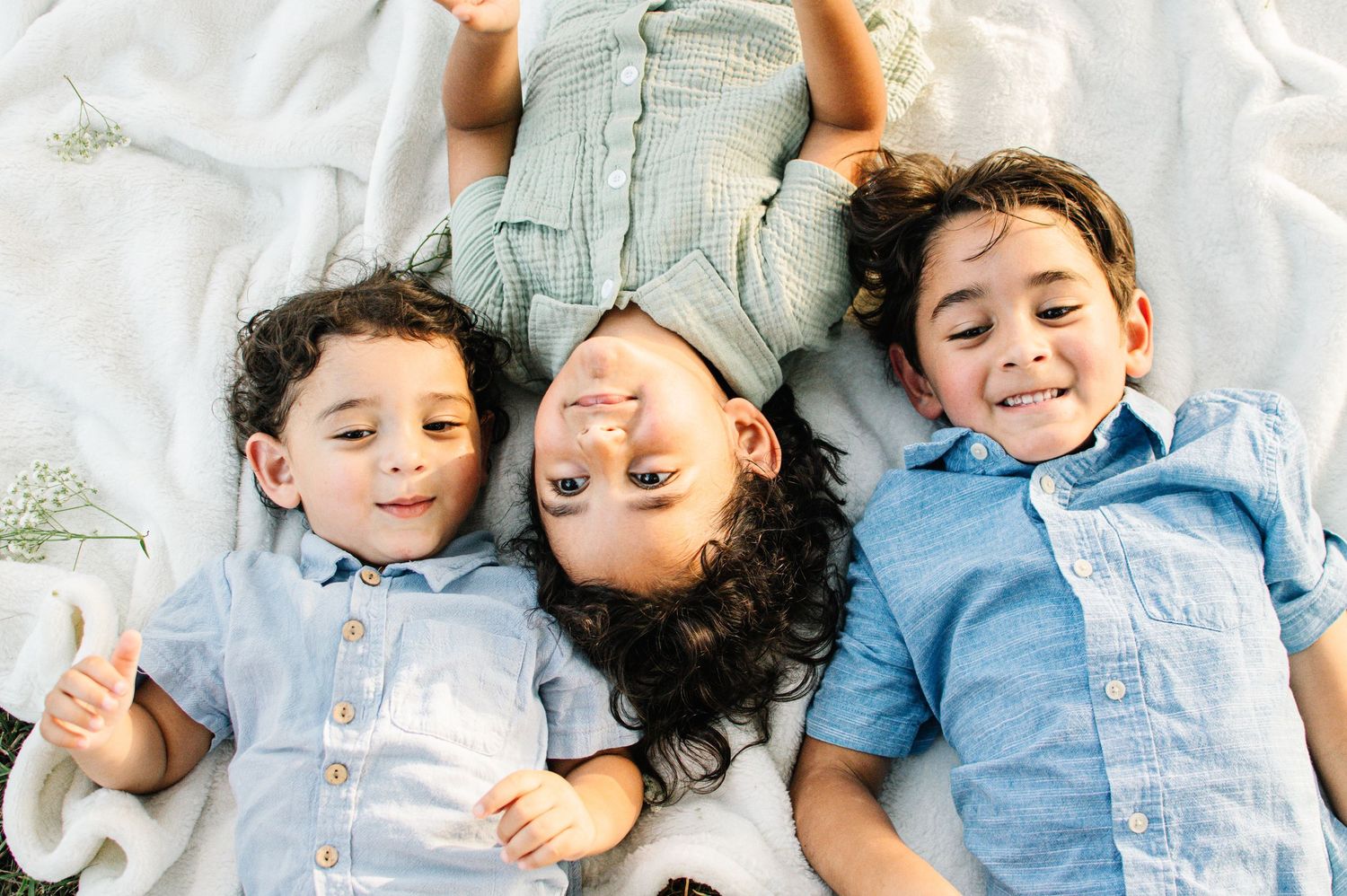 Three happy children lying on a white blanket outdoors, wearing matching light blue shirts, smiling and enjoying a sunny day.