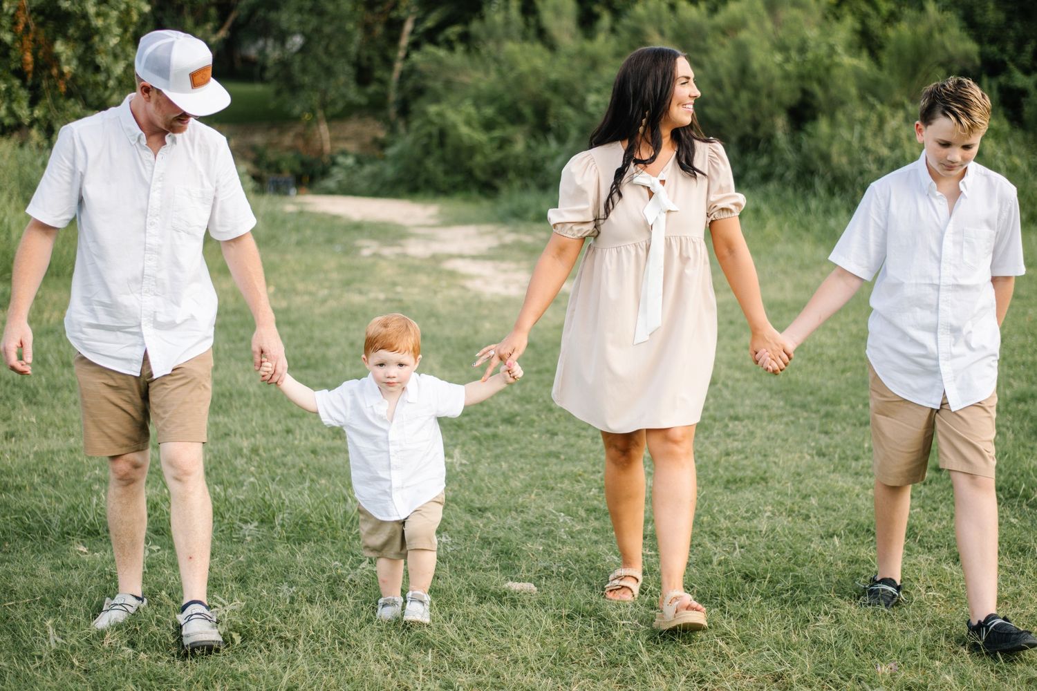 Family of four walking hand-in-hand through a grassy park in matching neutral tones on a sunny day.