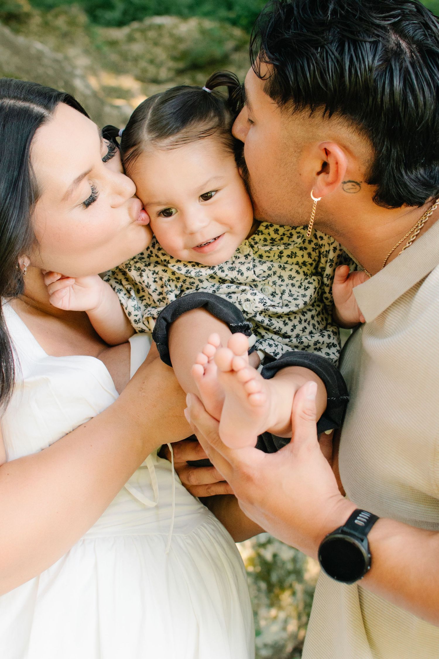 Parents kissing their smiling baby girl on the cheeks while holding her tiny feet outdoors.