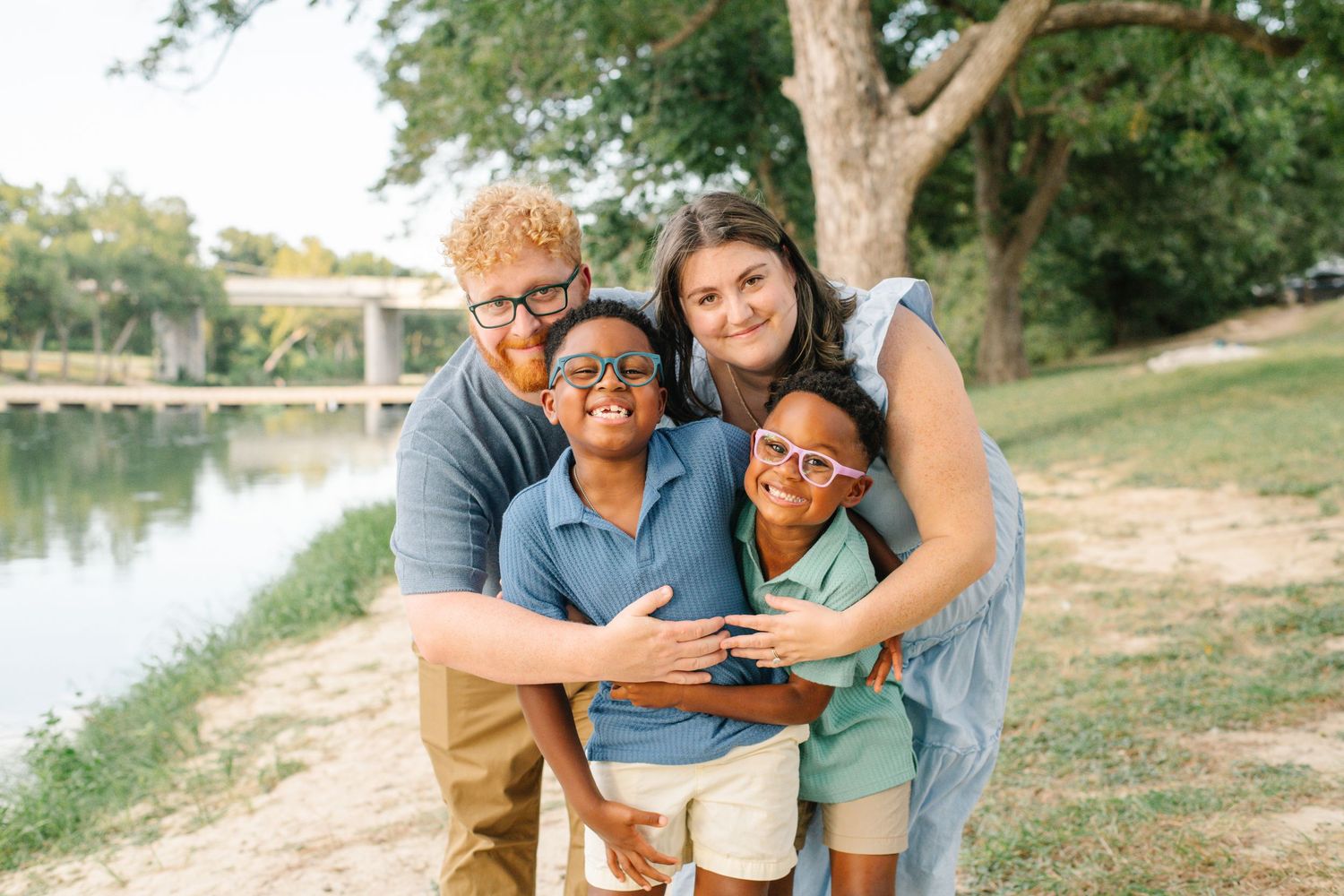 Happy family of four smiling outdoors near a river, with two children wearing colorful glasses on a sunny day.