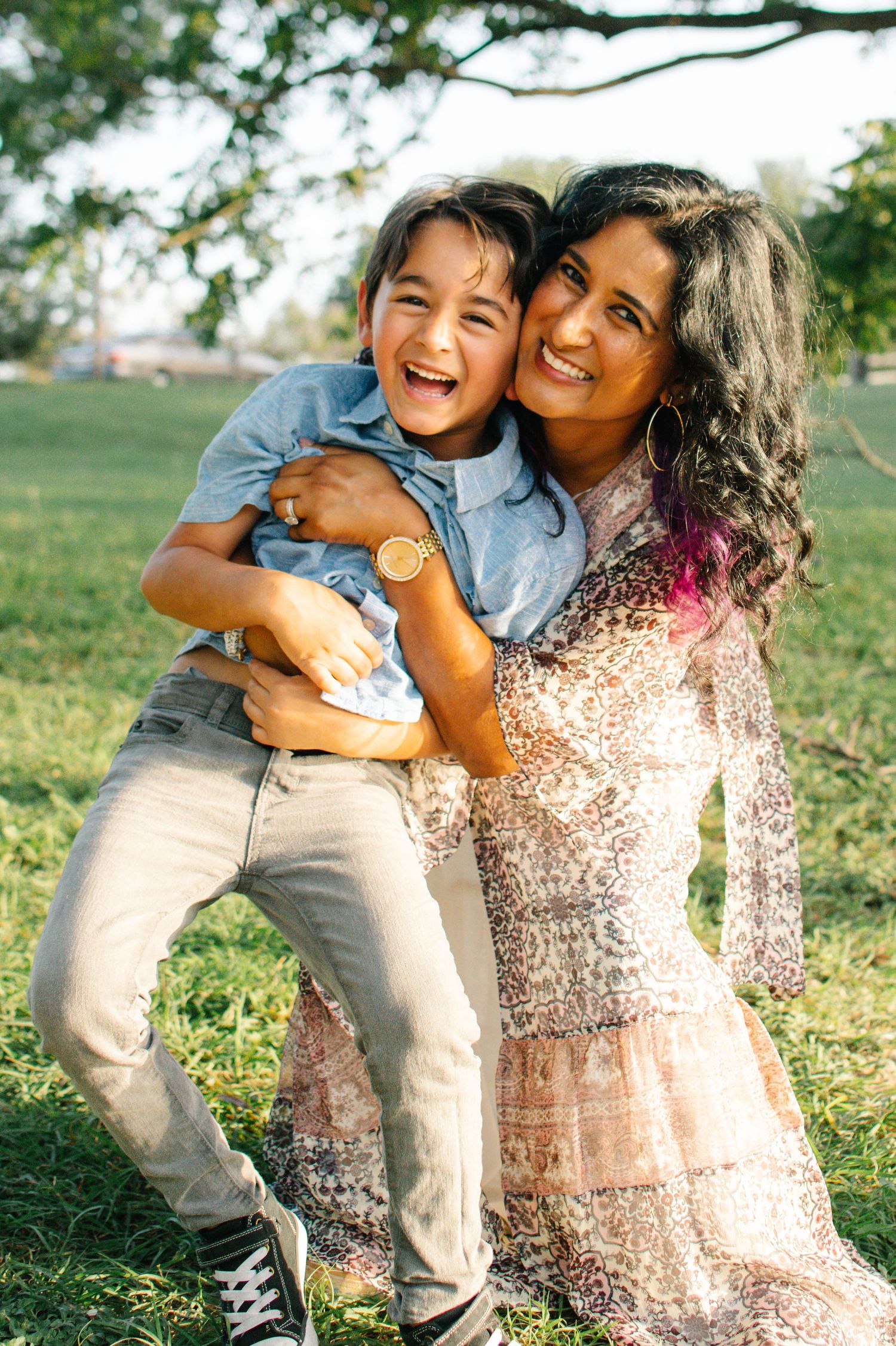 A joyful mother hugs her laughing young son outdoors in a sunny green park.