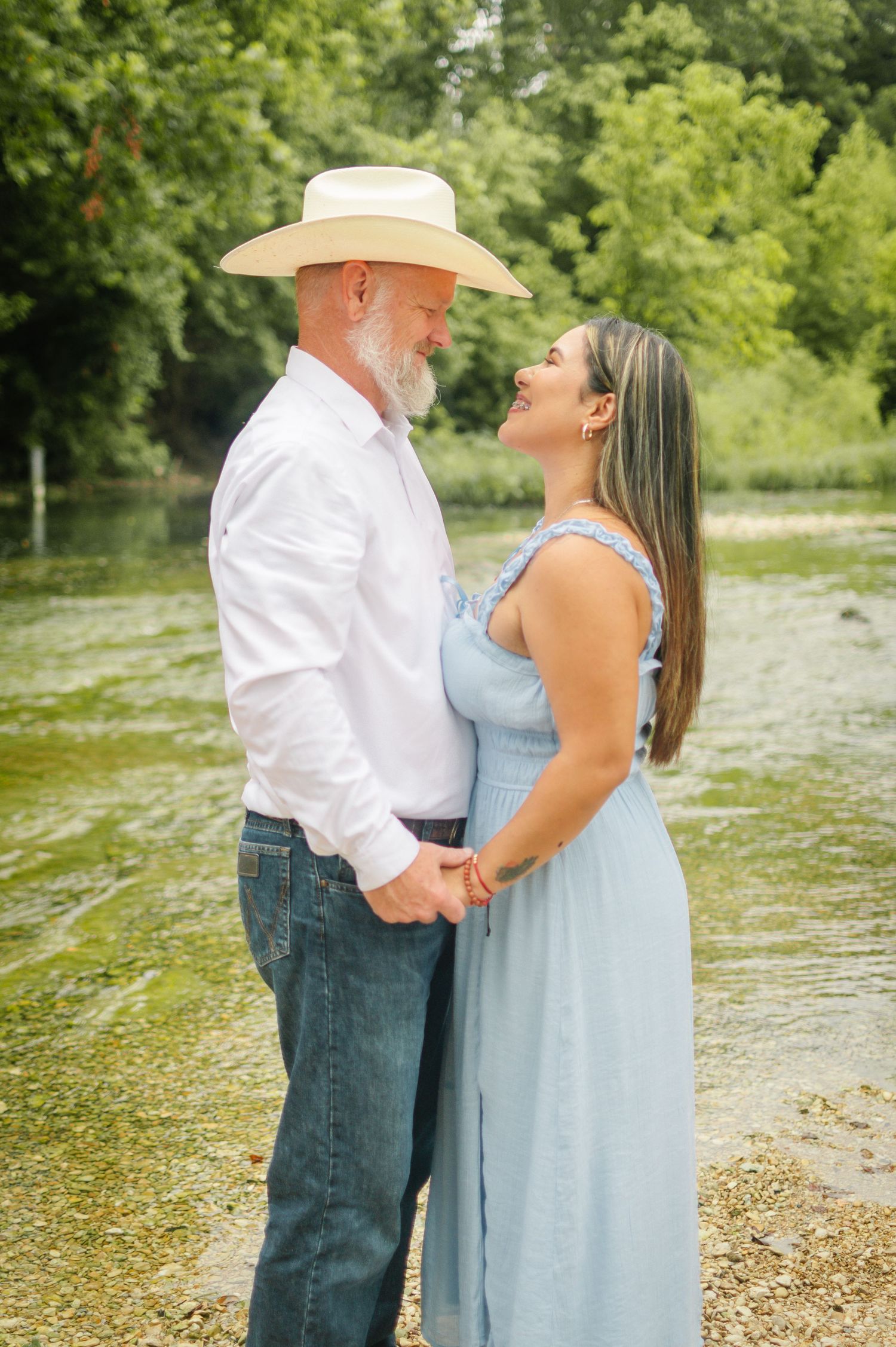Couple holding hands and smiling at each other by a river, man in cowboy hat and white shirt, woman in light blue dress.
