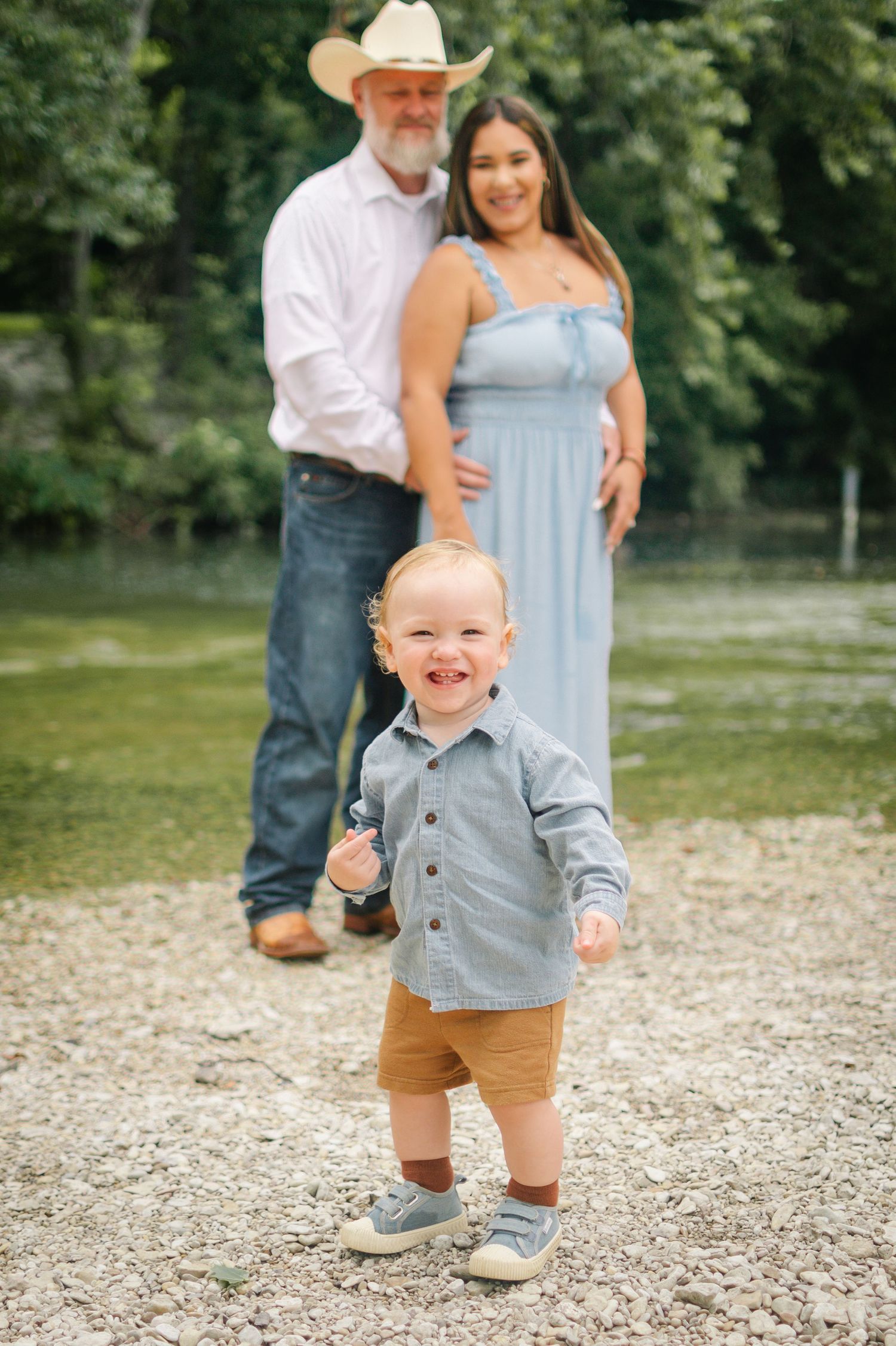 A smiling toddler boy stands on gravel while his parents pose together in a scenic outdoor family portrait.
