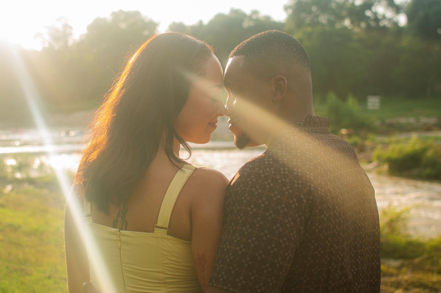 Couple sharing a romantic moment outdoors at golden hour with sunlight streaming through trees near a serene lake.