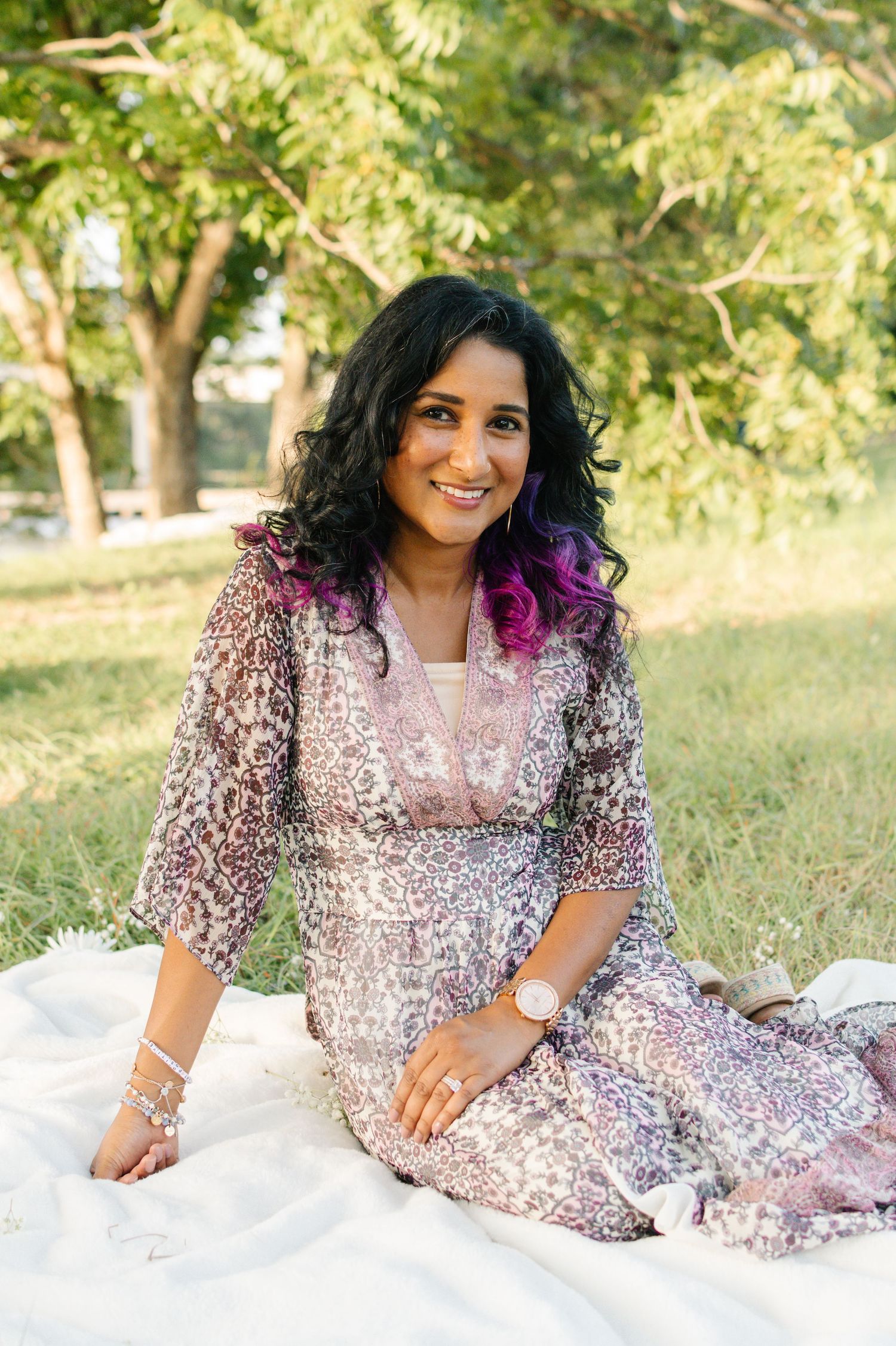 Smiling woman in floral dress sitting on white blanket in park with green trees.