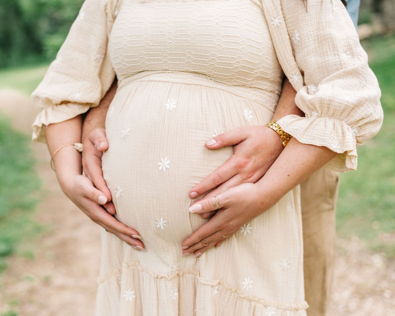 Pregnant woman in cream floral dress cradling baby bump outdoors, wearing gold bracelet.